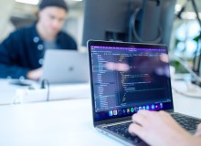 Ein Laptop steht auf einem Schreibtisch. Hinter diesem Laptop ist eine Person unscharf zu erkennen. Auf dem Display des Laptops befindet sich Code.