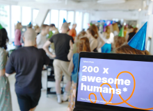 Im Vordergrund ist auf einem Laptopbildschirm zu lesen, dass mehr als 200 Mitarbeitende nun beim DigitalService arbeiten; im Hintergrund sind unscharf zahlreiche Mitarbeitende zu erkennen, die gemeinsam feiern