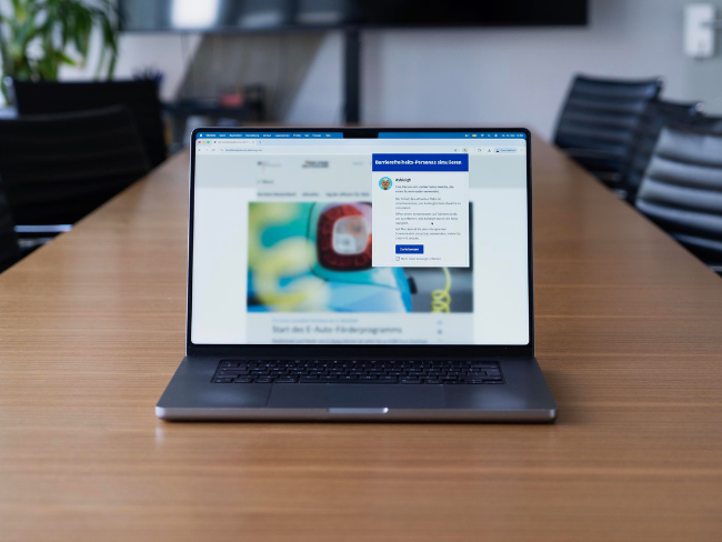 Laptop-Computer auf einem Tisch, der eine stark unscharf abgebildete Webseite in einem Web-Browser anzeigt mit einem Fenster darüber – zu einer stark seheingeschränkten Barrierefreiheits-Persona namens Ashleigh