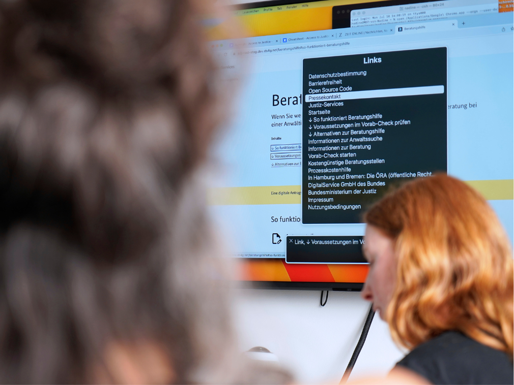 Eine Person sitzt vseitlich or einem großen Bildschirm auf dem ein Test zur Barrierefreiheit durchgeführt wird; im Vordergrund ist unscharf eine weitere Person zu erkennen