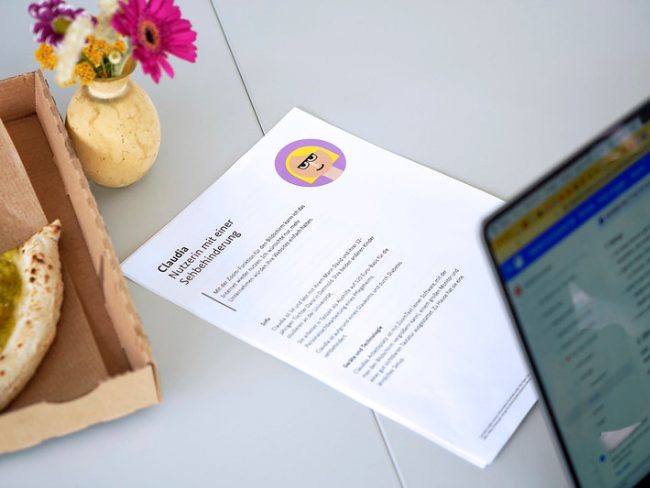 A table with a slice of pizza in a cardboard half, a small bouquet of flowers in a vase, a laptop with an open website and a printed text document with the title “Claudia – user with a visual impairment”