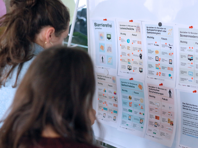 Two people stand in front of a whiteboard on which various pieces of paper are hung with instructions on the correct and incorrect design of accessible products