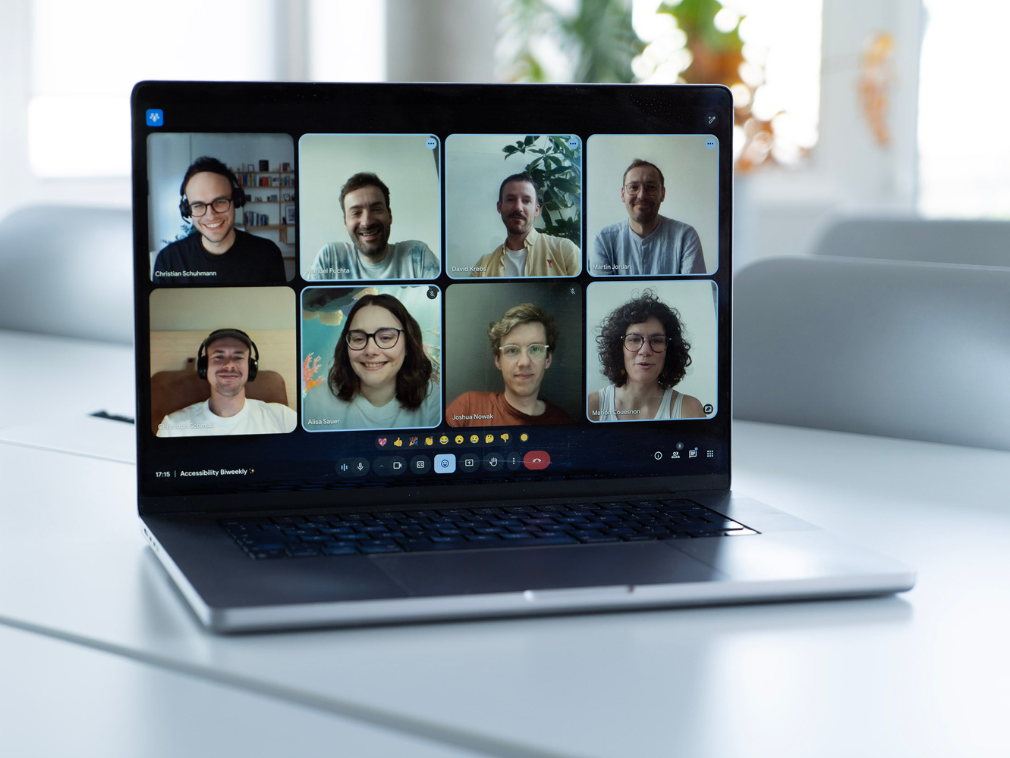 Ein Laptop steht auf einem Tisch in einem Büro; auf dem Bildschirm sind acht Personen in einem Video-Call zu sehen, die Teil der Arbeitsgruppe „Accessibility Ambassadors“ sind