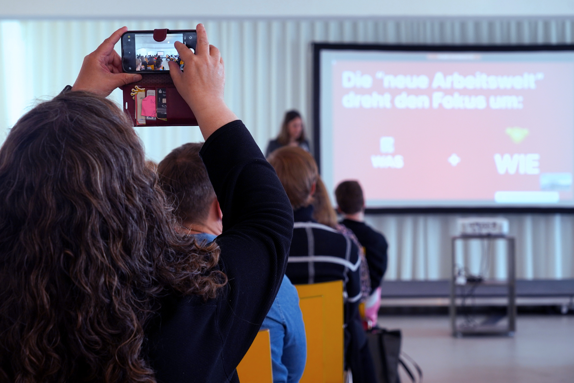 Der Rücken einer Person ist zu sehen. Sie sitzt in der vierten Sitzreihe bei einem Vortrag und filmt die vortragende Person mit ihrem Smartphone. Eine große Folie ist an die hintere Wand projiziert.