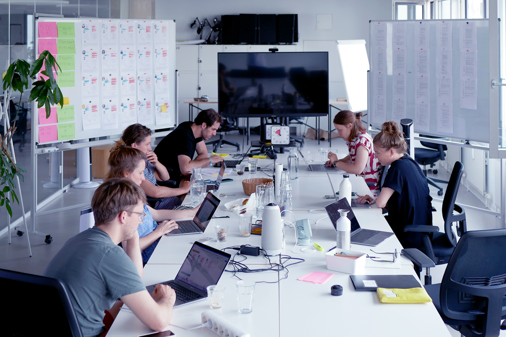 Sechs Personen sitzen an einem langen Tisch. Alle schauen konzentriert auf ihre Laptops. Links und rechts vom Tisch stehen große Whiteboards. An den Whiteboards sind bunte beschriebene Zettel und bedruckte DIN A4-Blätter angebracht.
