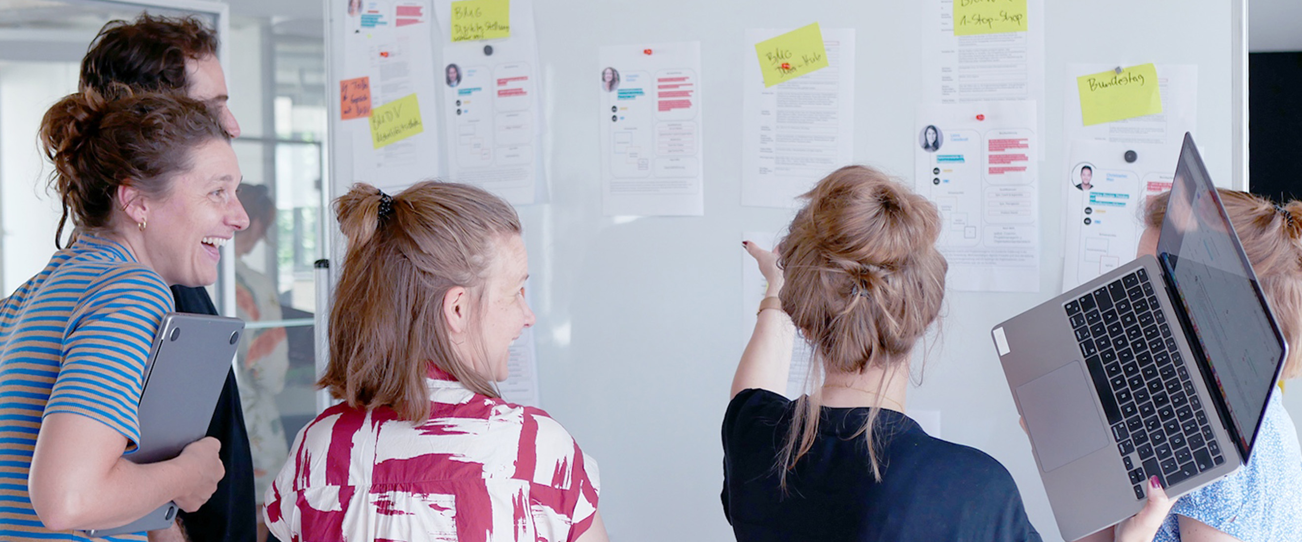 Fünf Personen stehen vor einem Whiteboard. Eine Person zeigt auf dieses. Die anderen sind der Person zugewandt. Die Stimmung wirkt locker und heiter. Auf dem Whiteboard hängen Zettel und bedruckte DIN A4-Blätter.