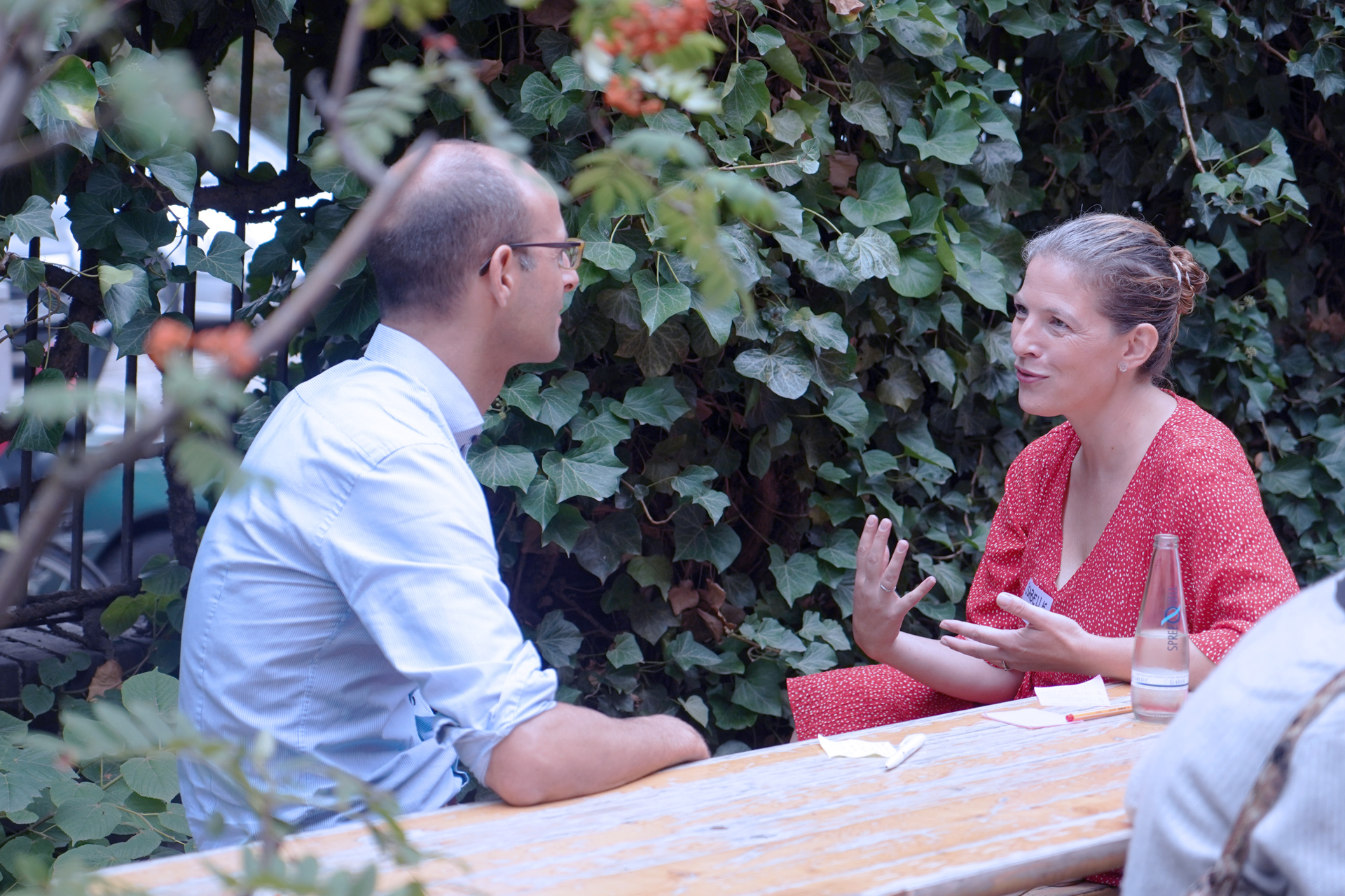 Zwei Personen unterhalten sich auf einer Bierbank sitzend. Eine Person gestikuliert. Im Hitergrund ist ein mit Efeu bewachsener Zaun.