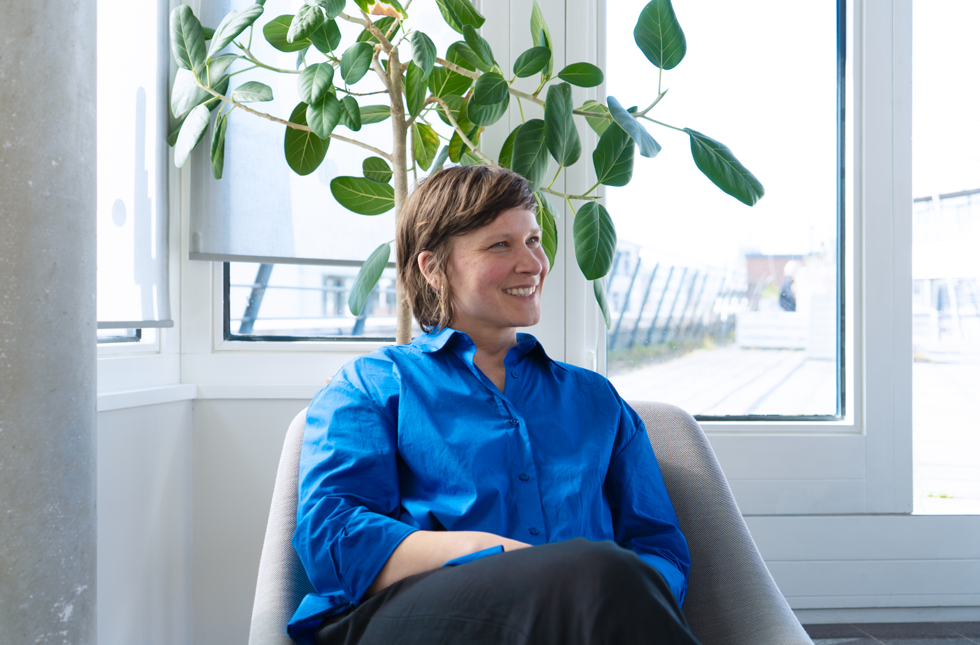 Hannah Bergmann sitzt auf einem Stuhl und lächelt eine Person an. Hinter ihr steht eine große grüne Pflanze, Licht fällt duch die geöffnete Tür der Dachterrasse.