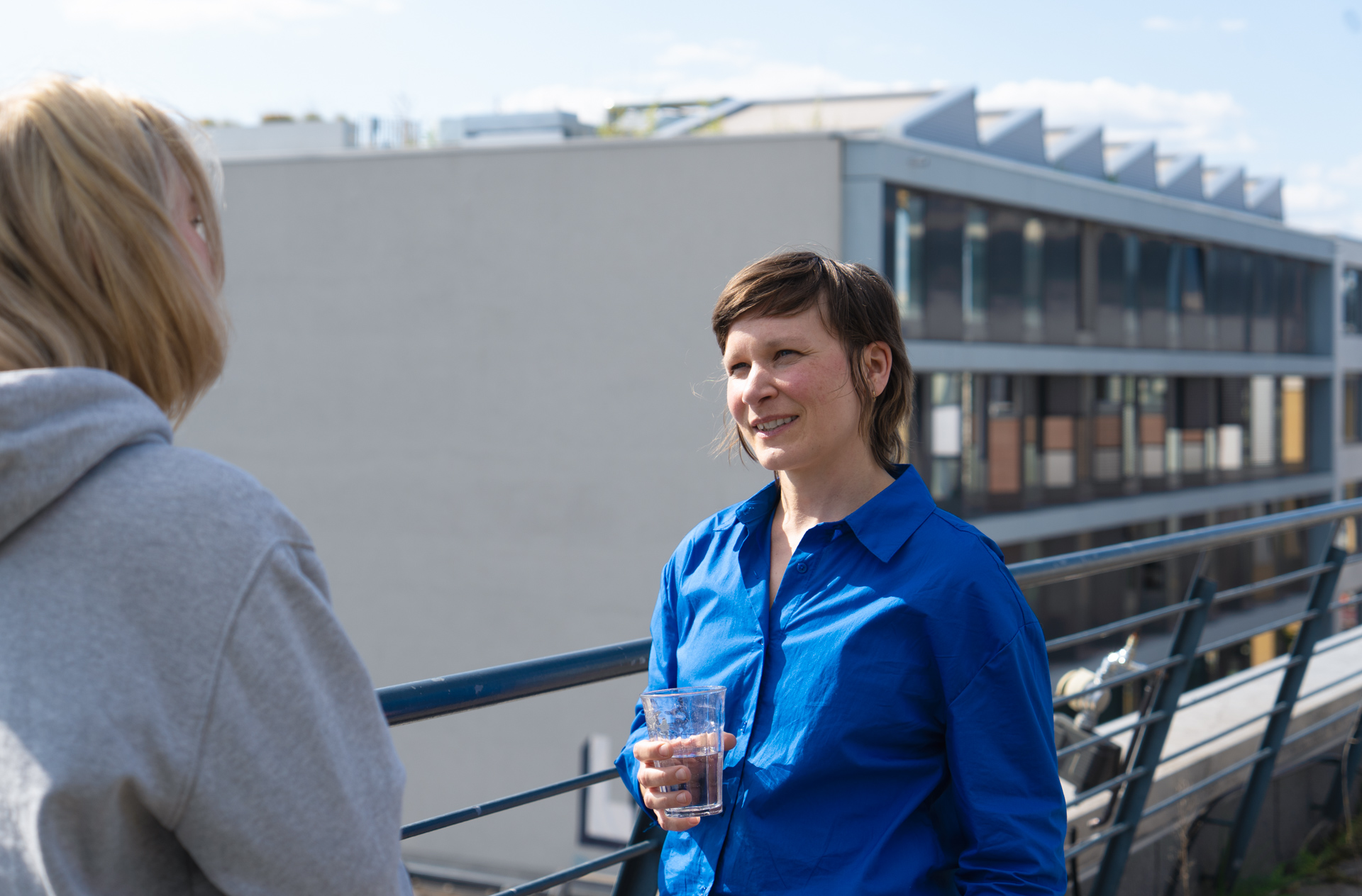 Hannah Bergmann steht auf der Dachterrasse des DigitalService. Sie hält ein Glas Wasser in der Hand und spricht mit einer Person die einen grauen Pullover trägt. Im Hintergrund ist eine Häuserfront zu sehen.