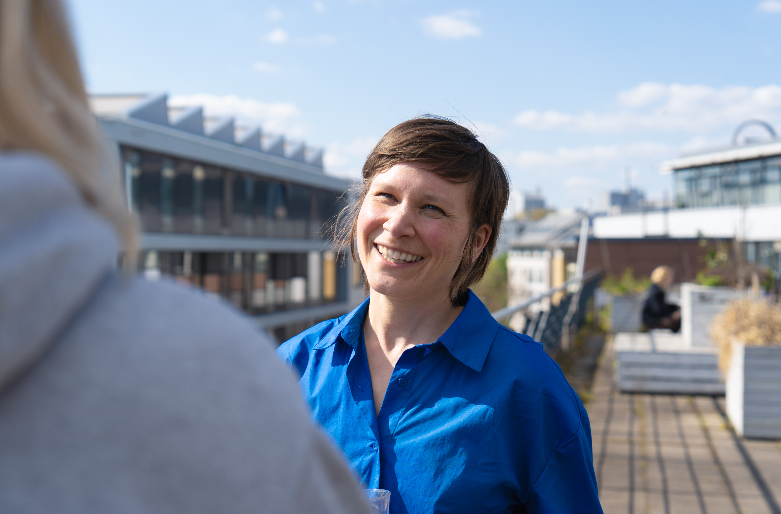 Hannah Bergmann steht auf der Dachterrasse des DigitalService und lächelt eine Person an. Im Hintergrund ist eine Sitzecke und mehrere Häuser zu sehen.