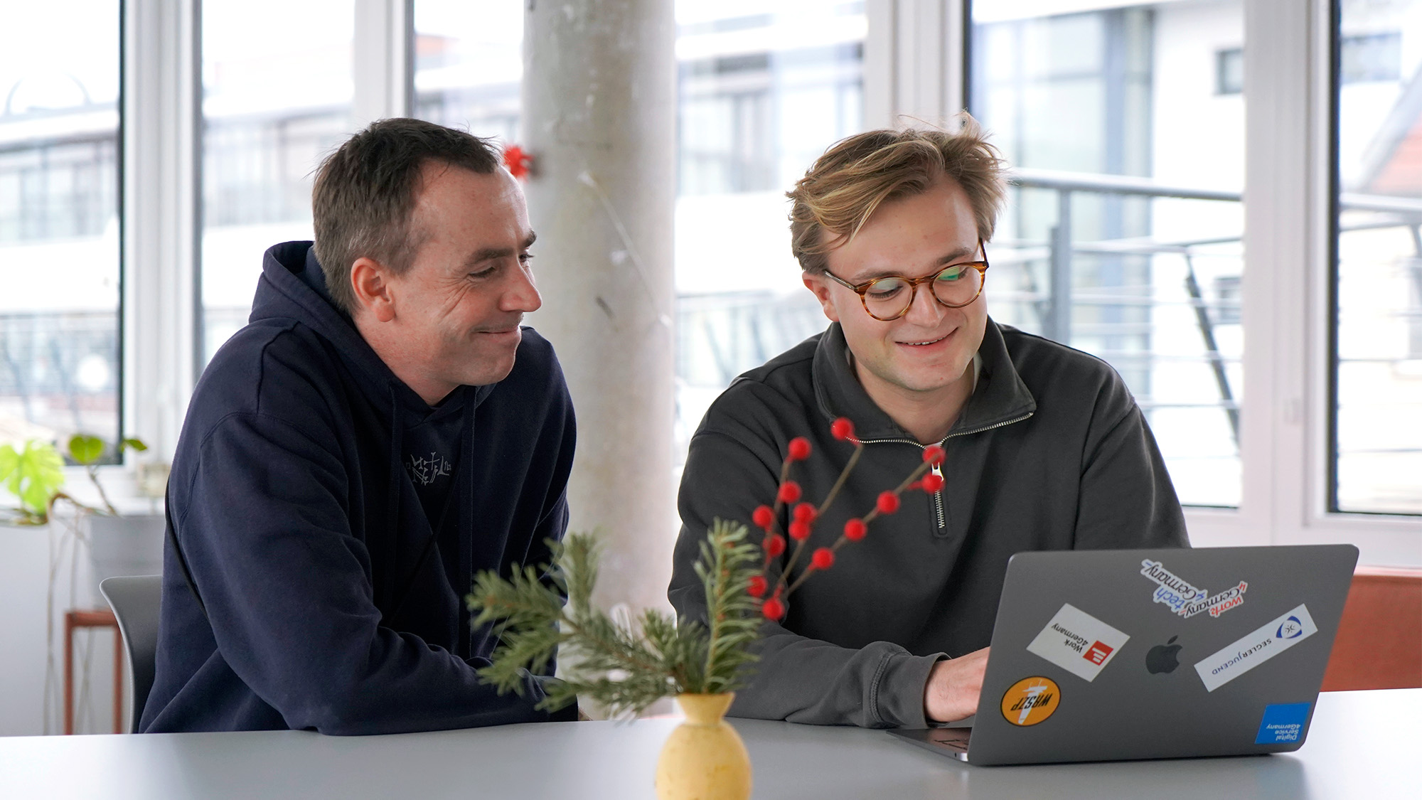 Interviewpartner Moritz im Gespräch mit einem Kollegen im Büro des DigitalService an einem Langen Tisch; beide schauen in das Laptop von Moritz