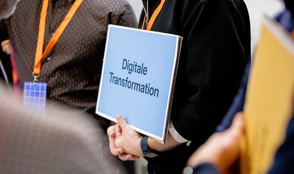 Person hält ein Schild mit der Aufschrift „Digitale Transformation“. Die Person trägt ein schwarzes Oberteil und ein orangefarbenes Lanyard. Weitere Personen mit Namensschildern sind im Hintergrund erkennbar.