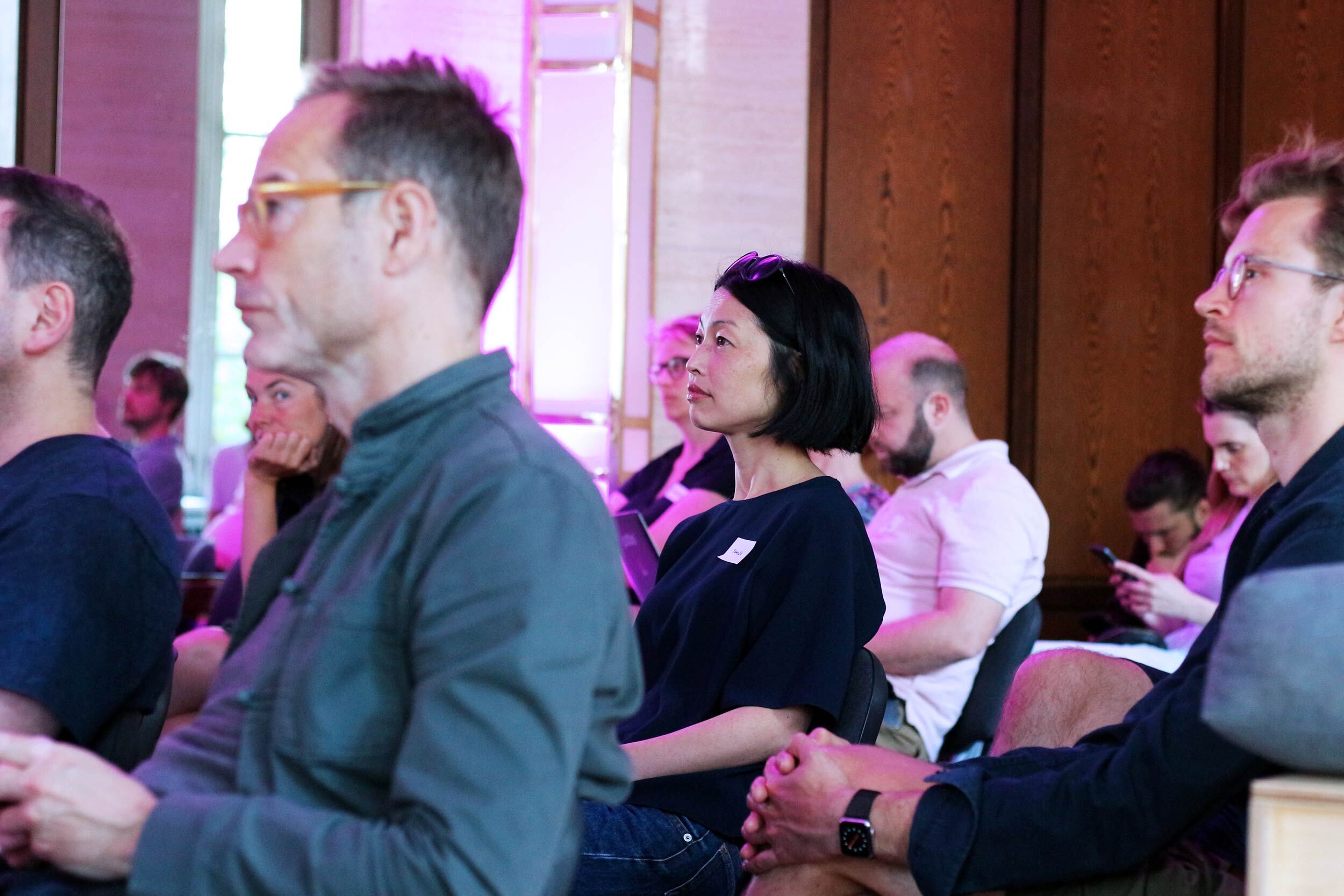 Knapp ein Dutzend Zuhörende verschiedener Altersgruppen und Ethnien sitzen in einem Raum mit Glas und Holzvertäfelung – im Zentrum des Bildes sitzt eine südostasiatische Frau mit Sonnenbrille im Haar