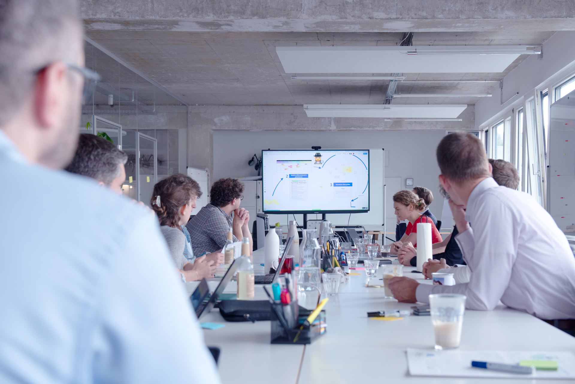 A larger group sits around a conference table and discusses; at the front end, a screen shows a presentation on the step-by-step development of the digital check.