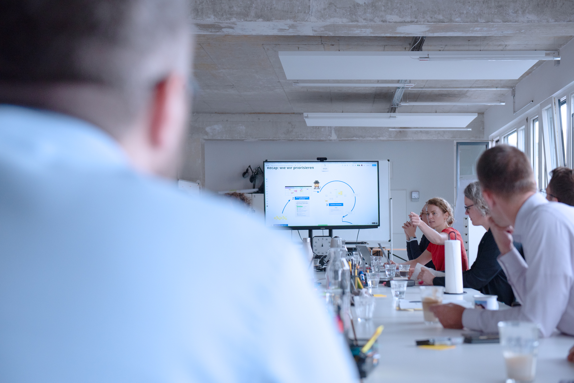 Eine größere Gruppe sitzt um einen Konferenztisch und diskutiert, am vorderen Ende ist auf einem Bildschirm eine Präsentation zur schrittweisen Weiterentwicklung des Digitalcheck zu sehen.