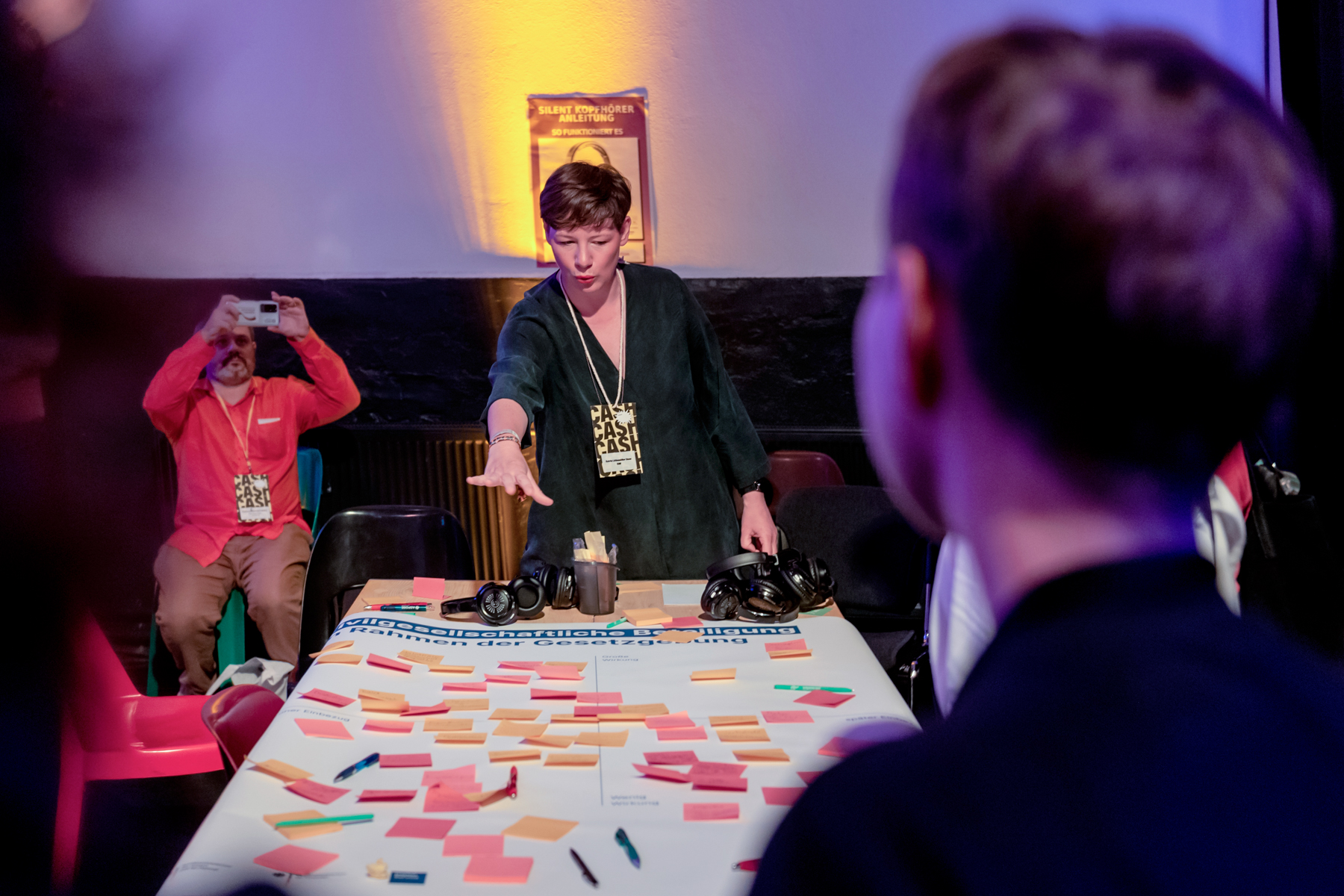 Auf einem Tisch liegt ein größeres Poster mit vielen Notizen auf bunten Post-its. Darum gruppieren sich einige Menschen, eine Person spricht und deutet auf einen Bereich des Posters.