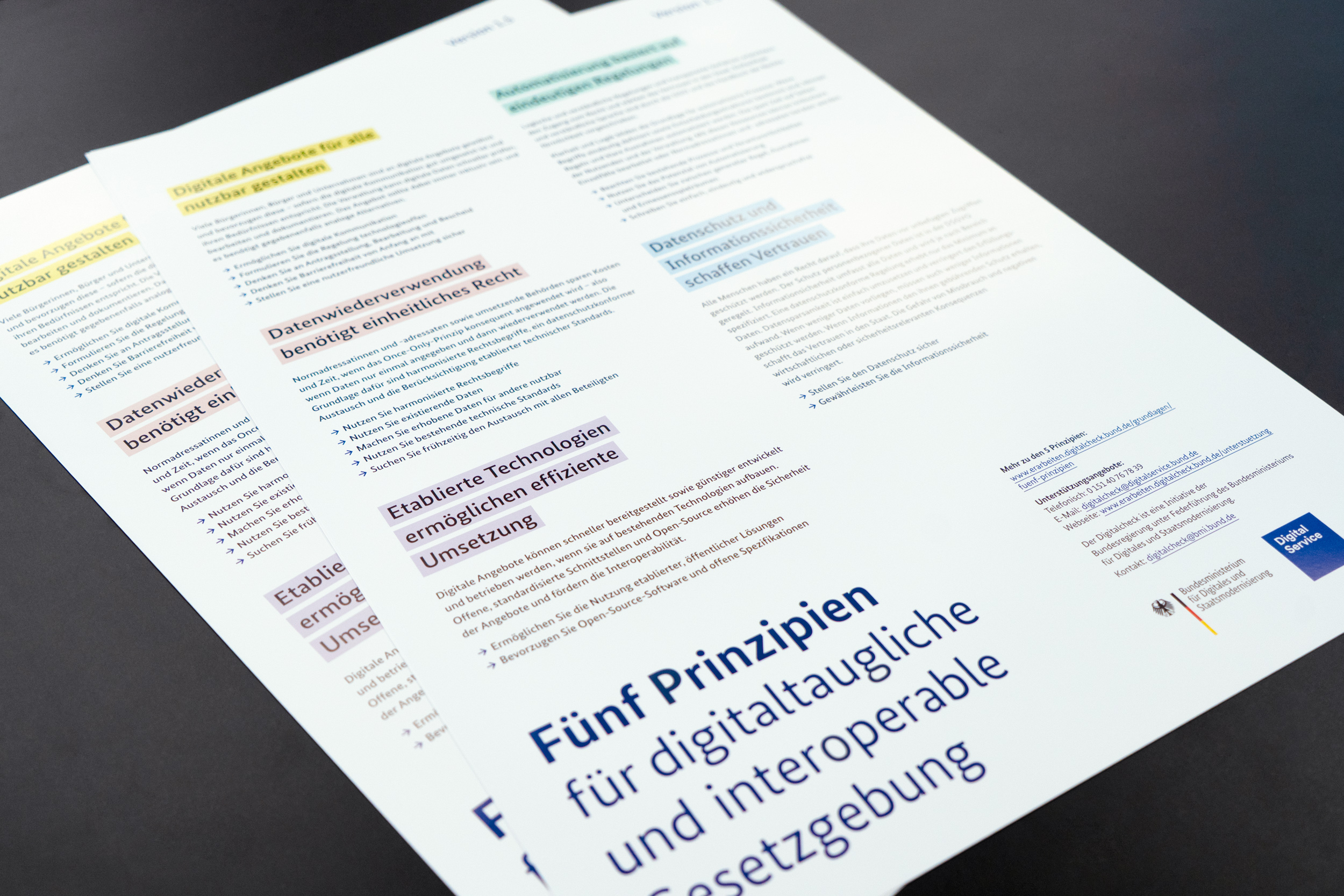 Zwei Poster mit den fünf Prinzipien für digitaltaugliches Recht liegen auf einem schwarzen Tisch. Die Texte auf dem Poster sind in verschiedenen Farben hervorgehoben, um unterschiedliche Prinzipien zu kennzeichnen. Unten rechts sind die Logos des Bundesministeriums für Digitales und Staatsmodernisierung sowie vom DigitalService zu sehen.