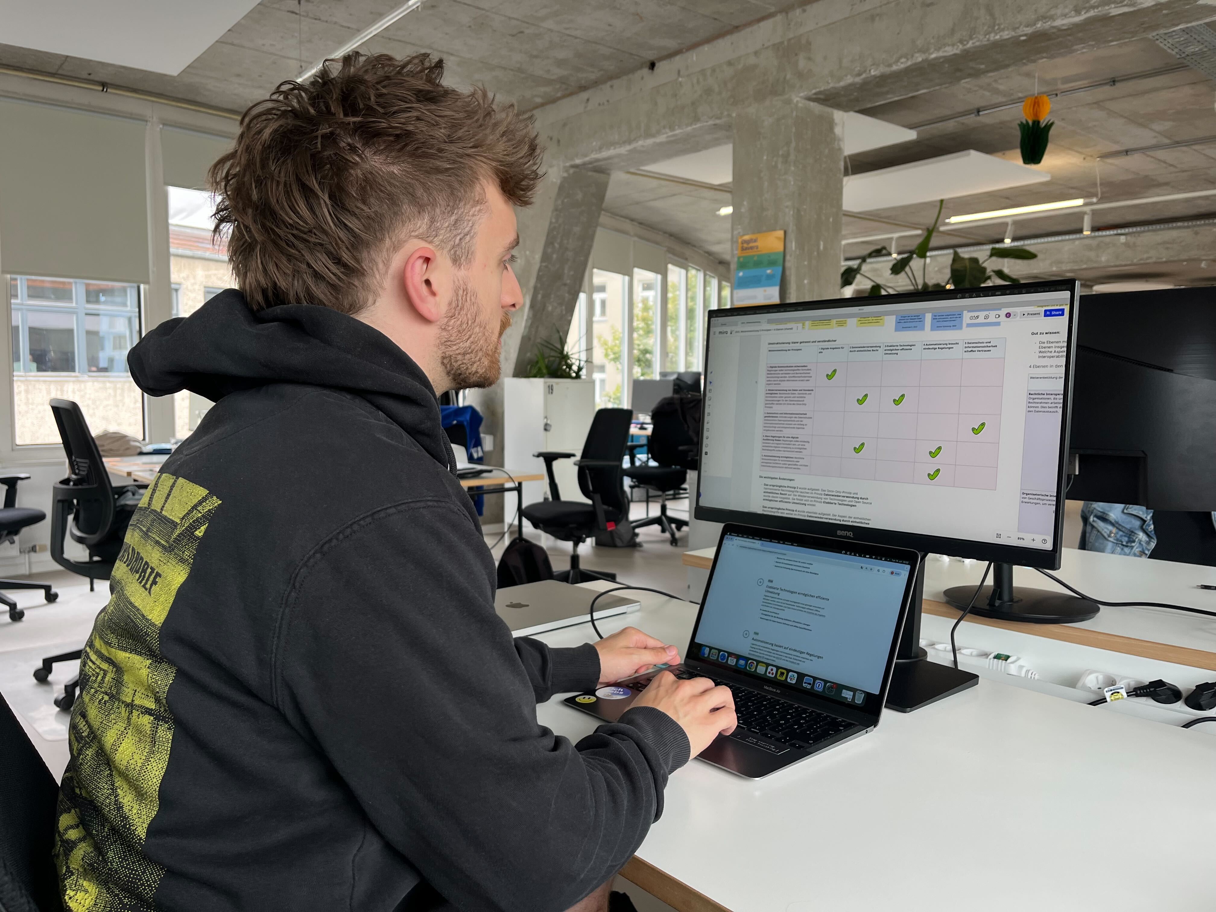 Hendrik Schmidt sitzt an einem Schreibtisch und arbeitet an einem Laptop, der mit einem großen externen Monitor verbunden ist. Auf dem Monitor ist eine Tabelle mit den alten und neuen Prinzipien für digitaltaugliches Recht zu sehen.