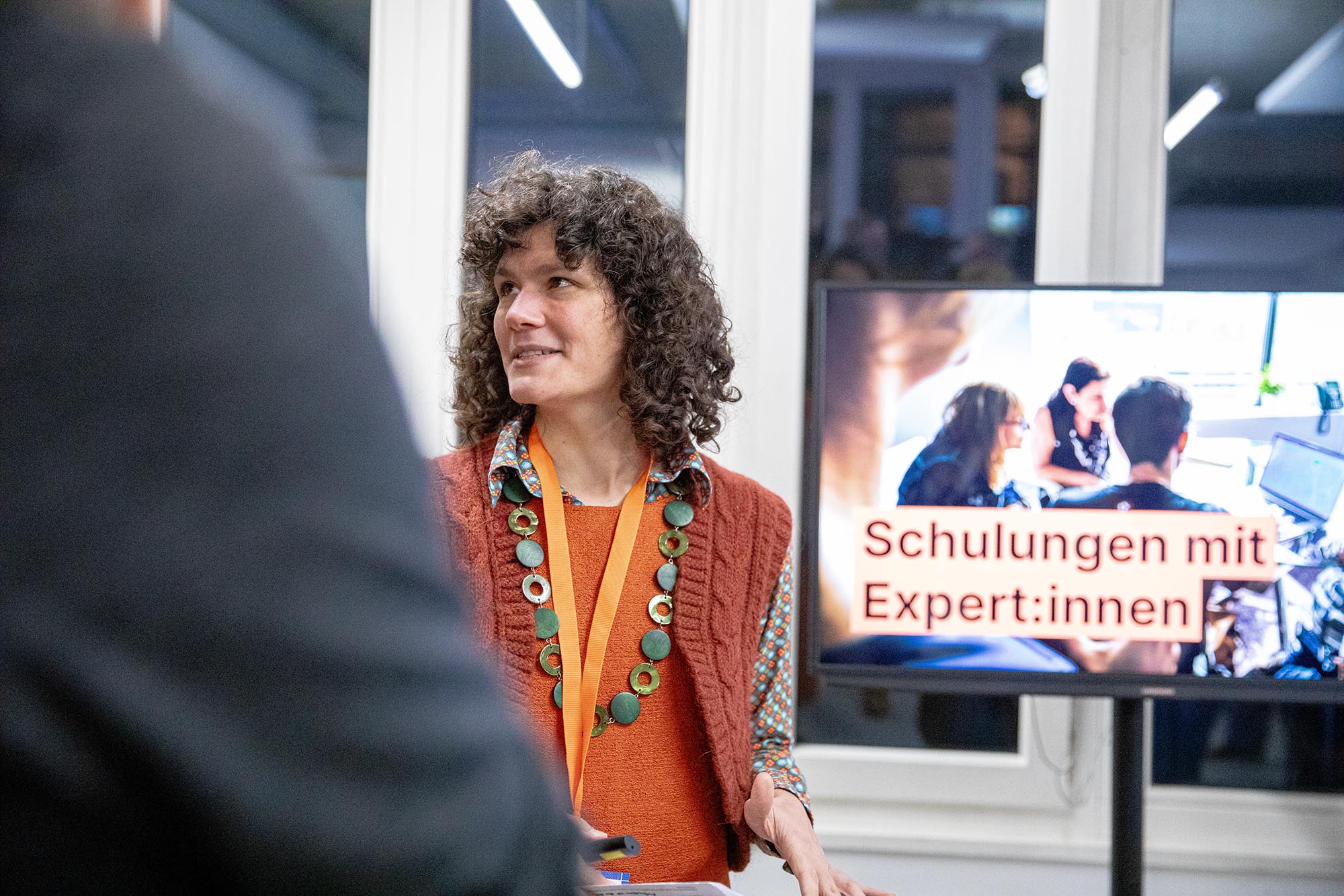Autorin Marion bei einer Präsentation ihrer Arbeit neben einem Monitor, auf dem ein Foto von vier Menschen vor einem Laptop mit dem Titel „Schulungen mit Expert:innen“ zu sehen ist
