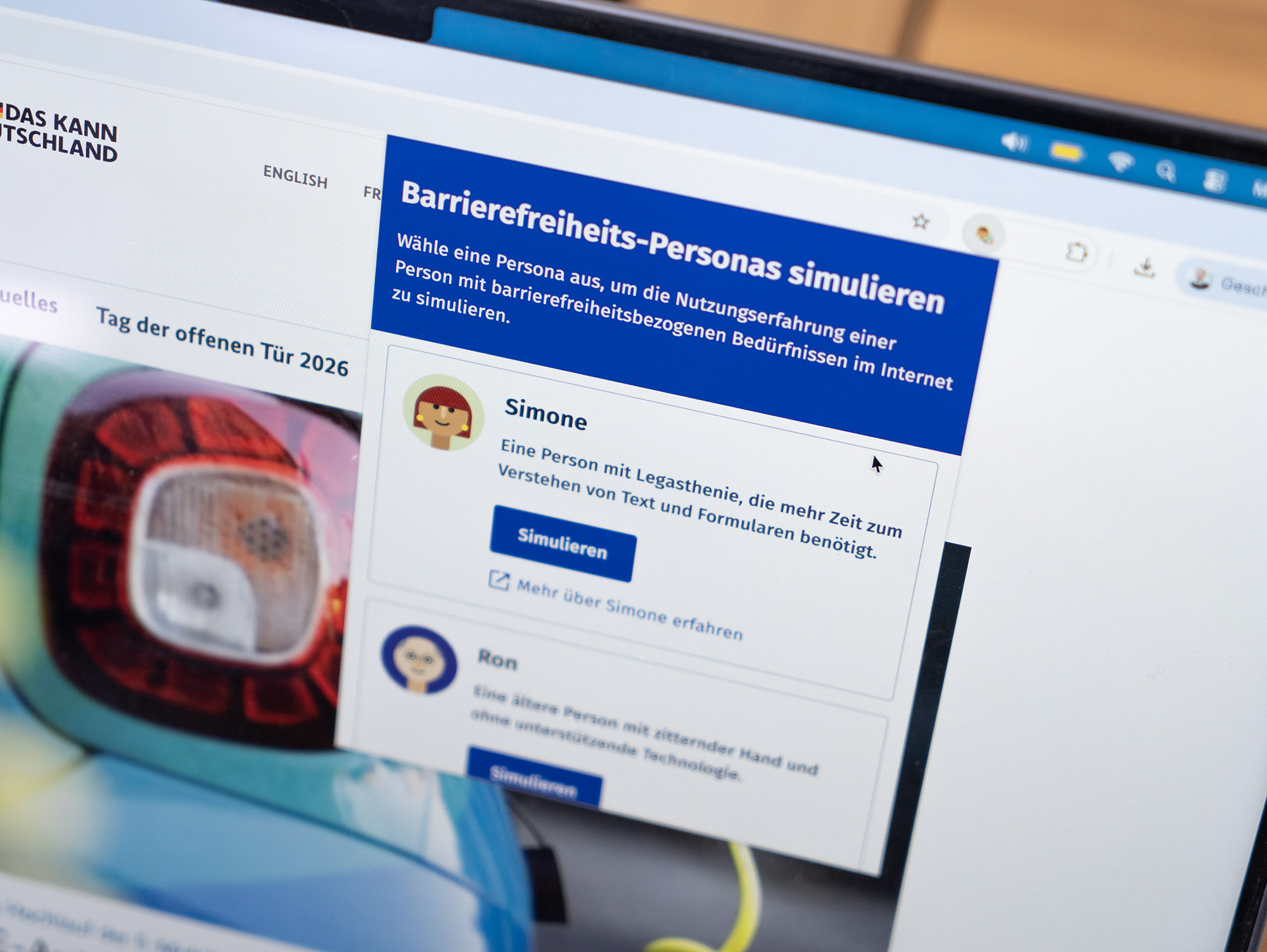 Auf dem Bildschirm eines Laptops wird eine Webseite mit einem Popup-Fenster mit der Überschrift „Barrierefreiheits-Personas simulieren“ angezeigt. Das Popup-Fenster zeigt 2 Optionen mit Simone und Ron, eine Person mit Legasthenie und zitternder Hand und ohne unterstützender Technologie, beide zeigen einen Knopf namens „Simulieren“