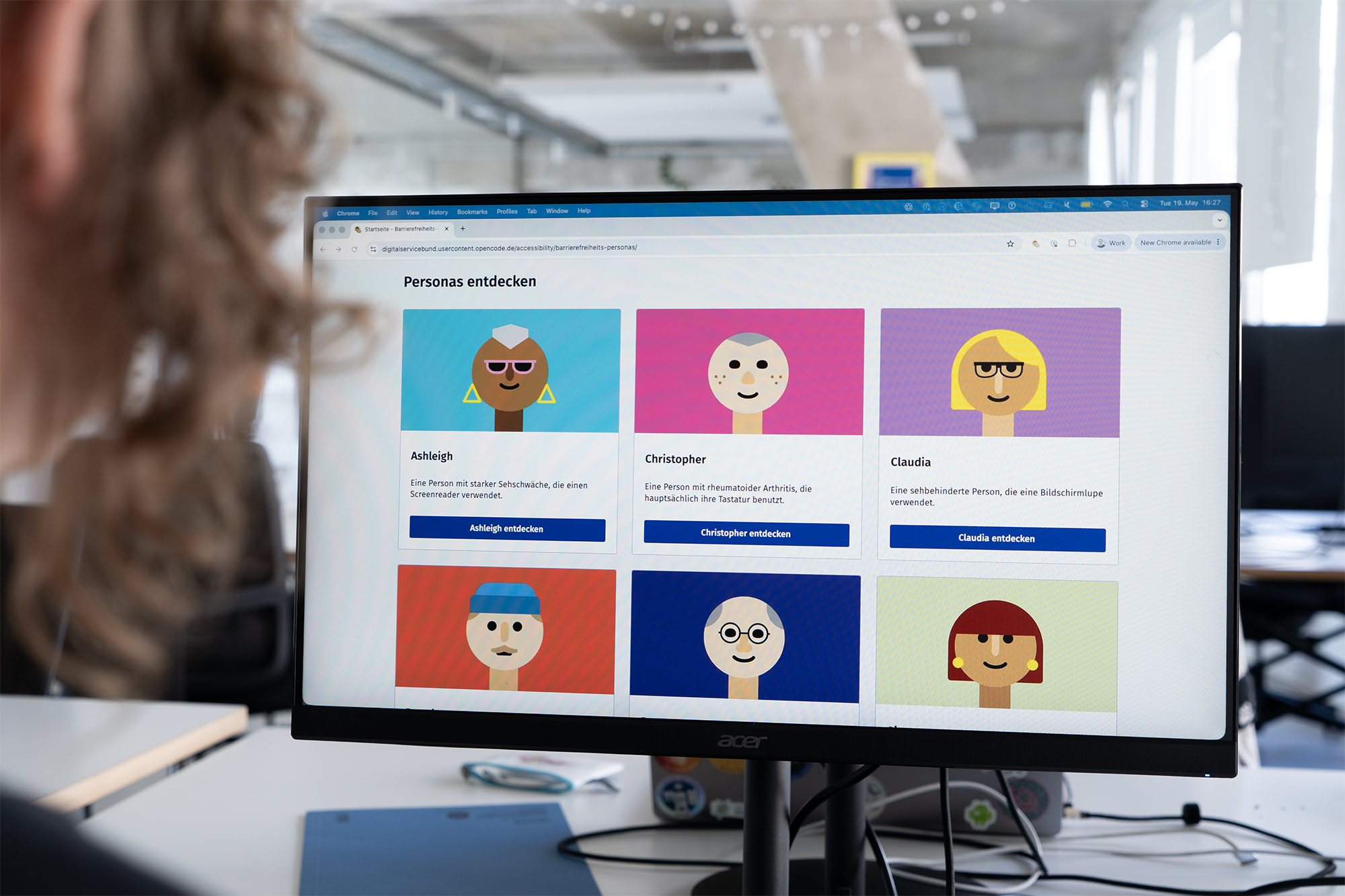 Computer-Monitor in einem modernen Büroumfeld, das 6 simple und farbenfrohe Illustrationen von Personen zeigt mit der Überschrift „Personas entdecken“, sie haben verschiedene Hauttöne, Frisuren, Accessoires und Alter, eine Person schaut auf den Monitor