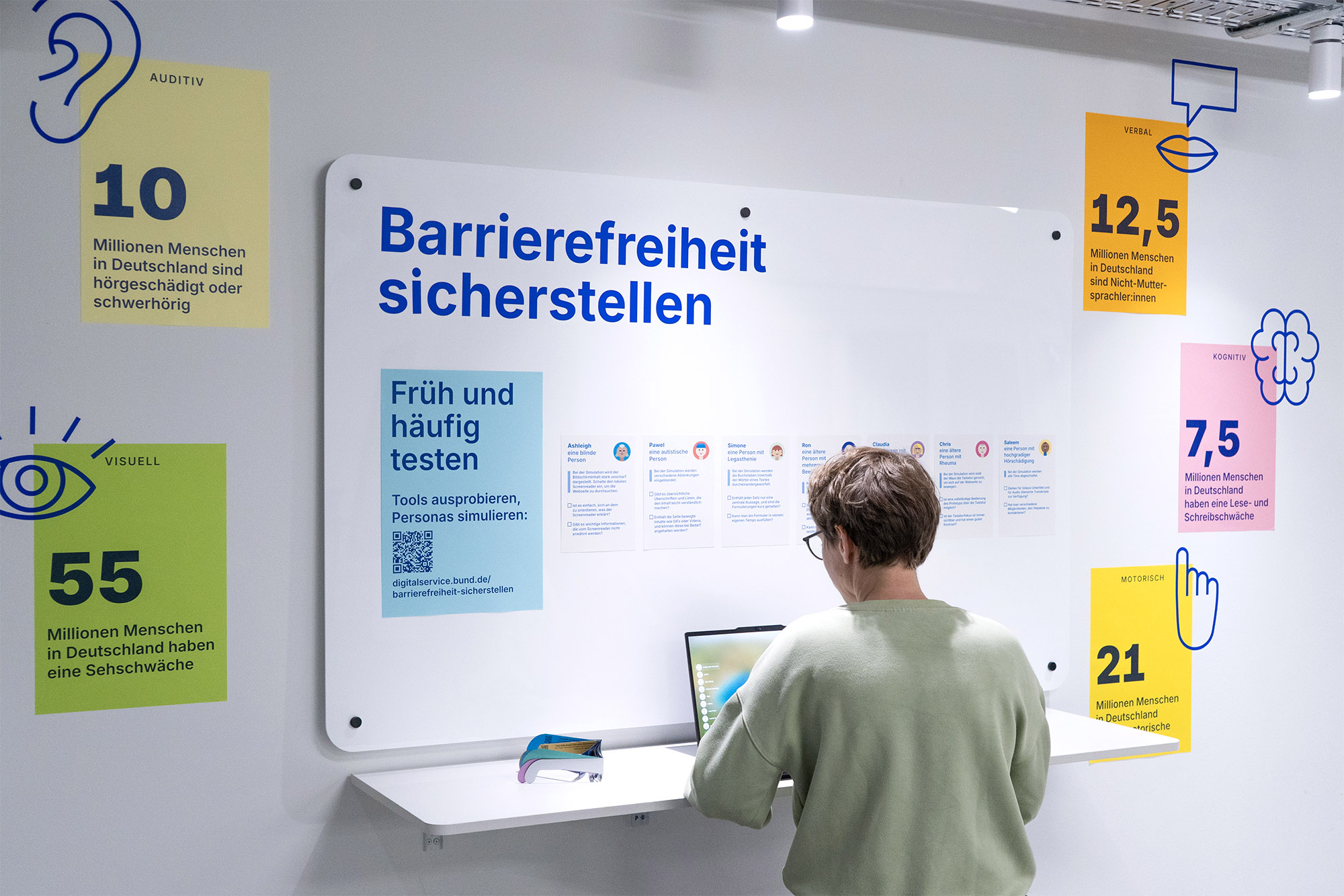 Eine Person sitzt an einer weißen Theke und beschäftigt sich mit an einem Laptop-Computer mit Thema Barrierefreiheit. Umgebende Poster und Icons zeigen Statistiken zu Behinderungen in Deutschland, darunter Seh-, Hör-, motorische, kognitive und sprachliche Beeinträchtigungen.