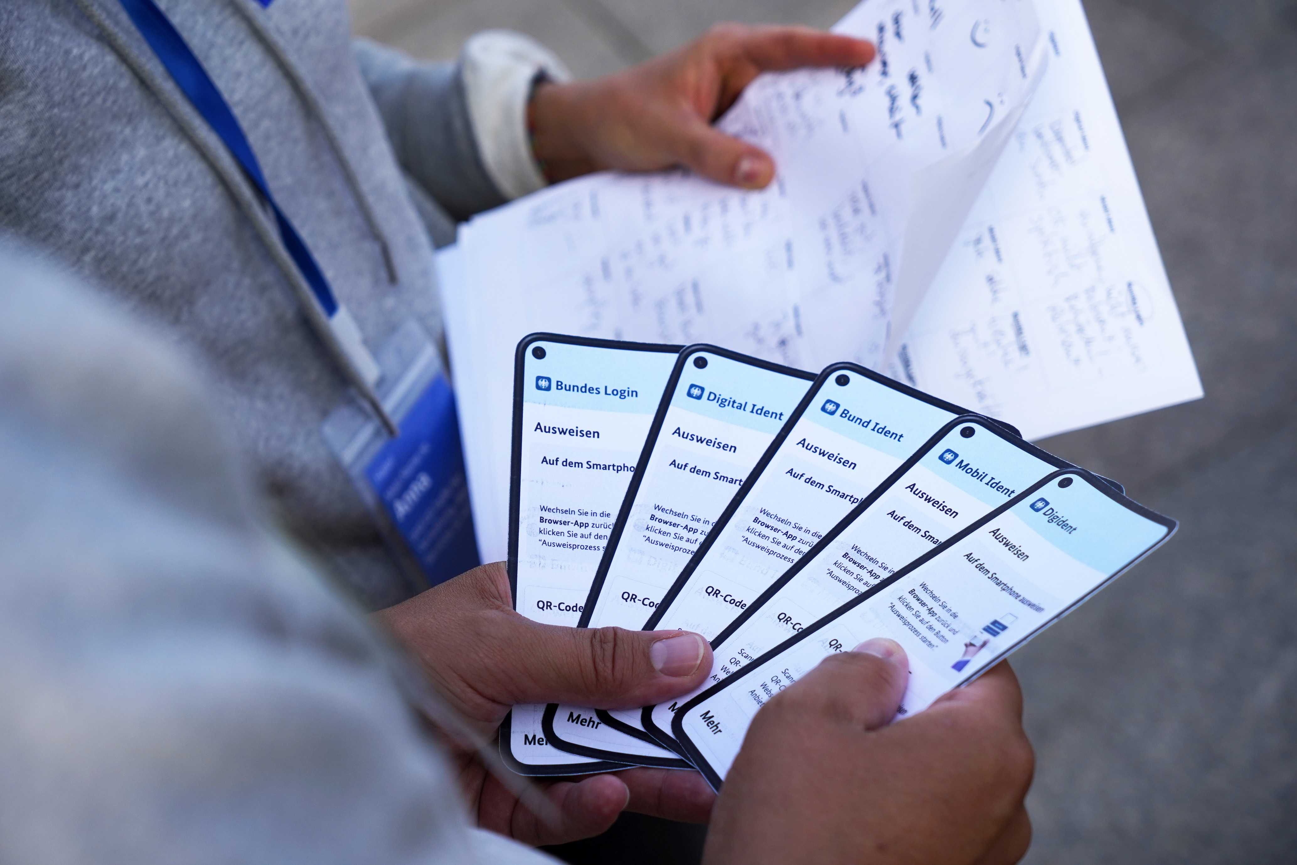 Eine Person hält fünf Papierkarten in Handy-Format mit unterschiedlich gestalteten Ansichten der BundesIdent App  in den Händen; eine zweite Person hält Zettel mit handschriftlichen Notizen in den Händen