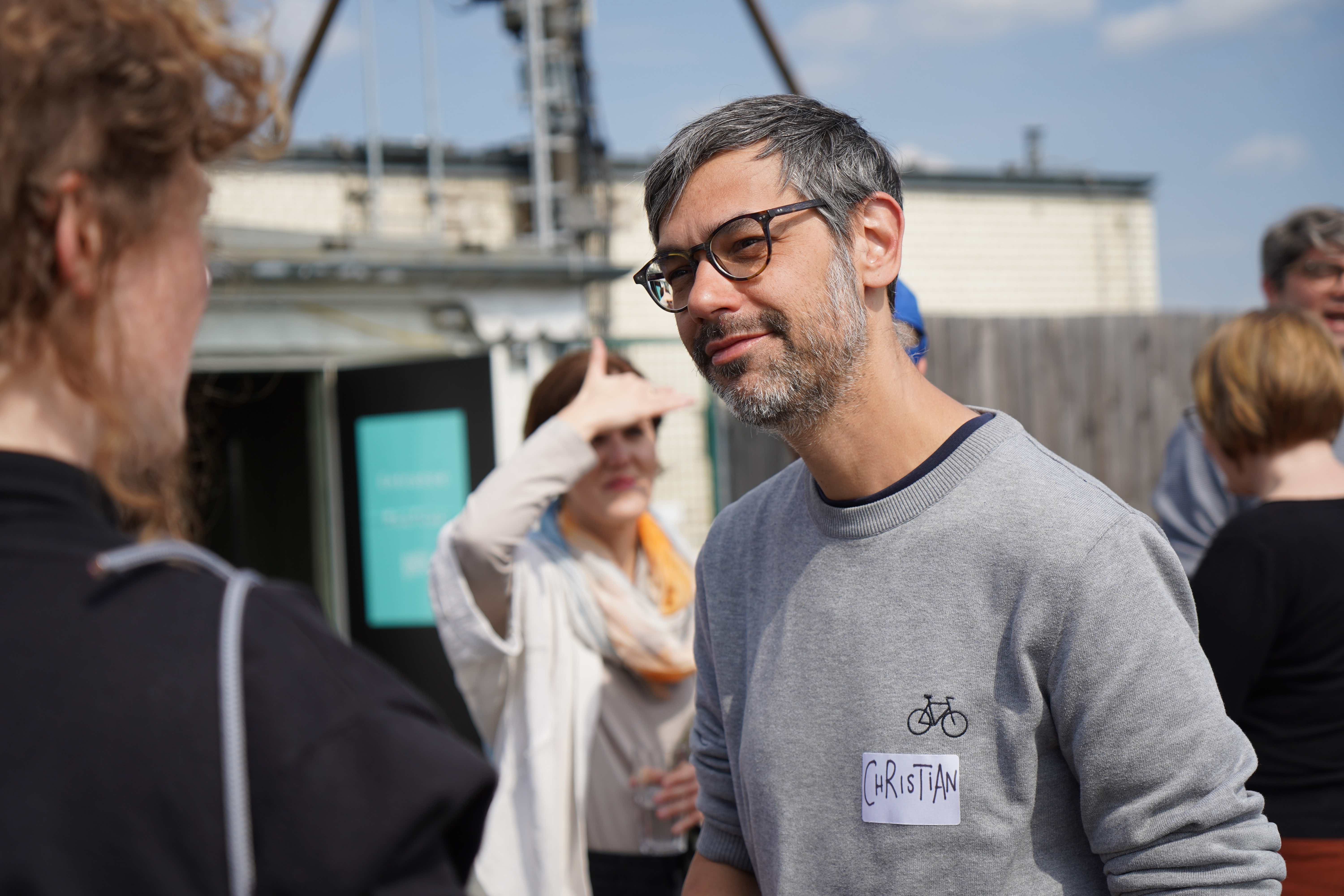 Portraitaufnahme von Christian Müller. Er unterhält sich konzentriert mit einer Gesprächspartnerin. Beide stehen draußen auf einer Terasse. Die Sonne scheint ihm ins Gesicht