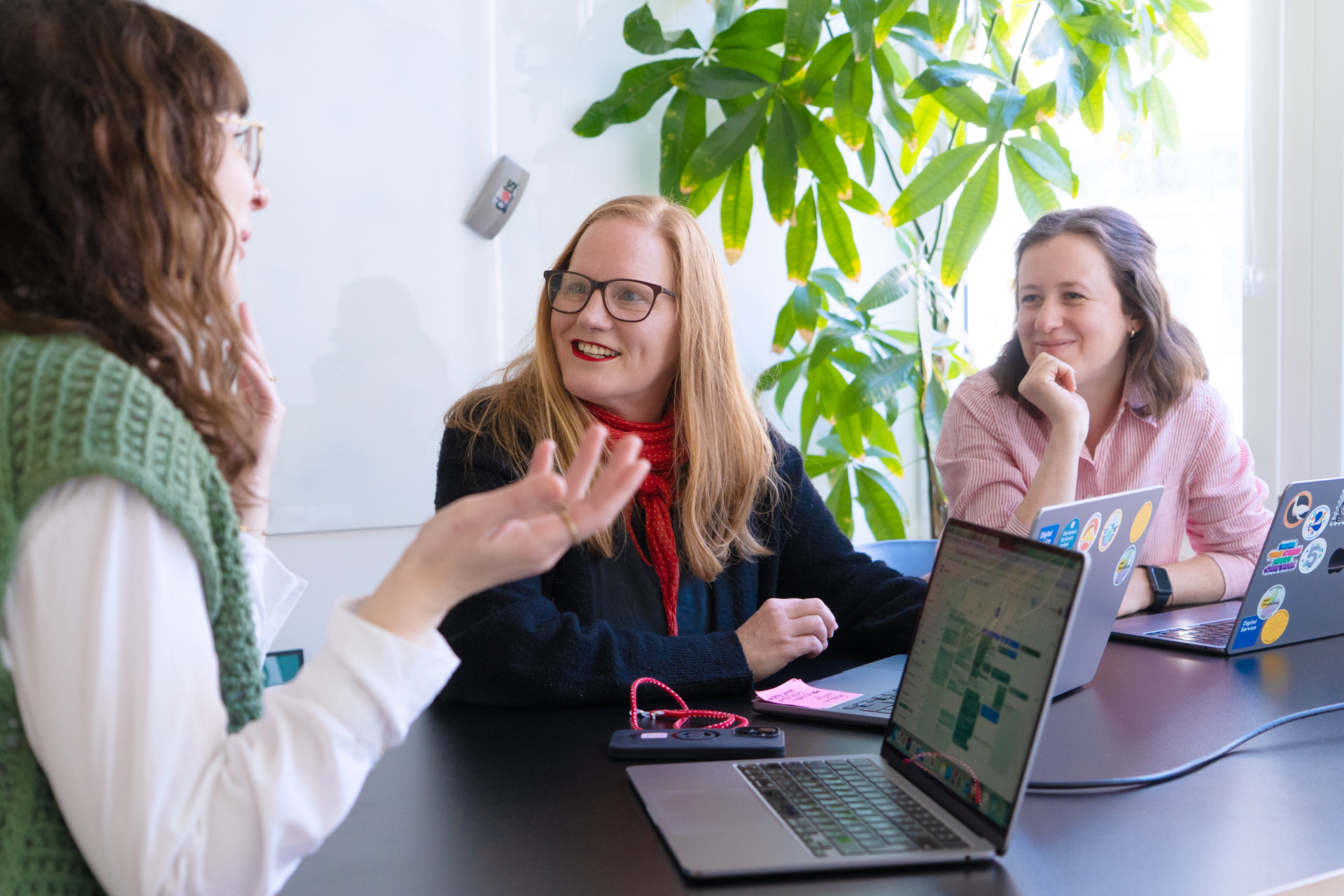 Mehrere Personen sitzen an einem Tisch in einem modernen Büroraum und sprechen angeregt miteinander. Auf dem Tisch stehen aufgeklappte Laptops.