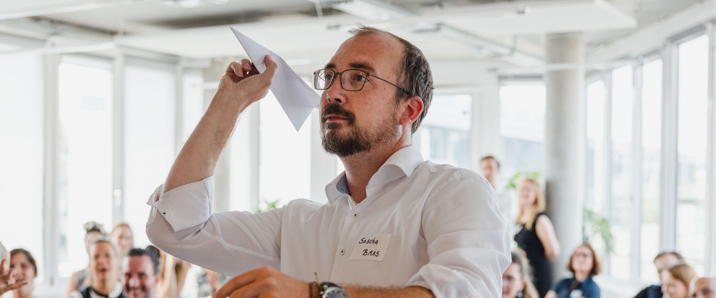 Projektpartner Sascha steht in einem Raum mit einem Papierflieger in der rechten Hand, bereit ihn losfliegen zu lassen. Im Hintergrund sind moderne Büroräume mit großen Fenstern zu sehen. Viele Menschen sitzen auf Stühlen und schauen Sascha an.