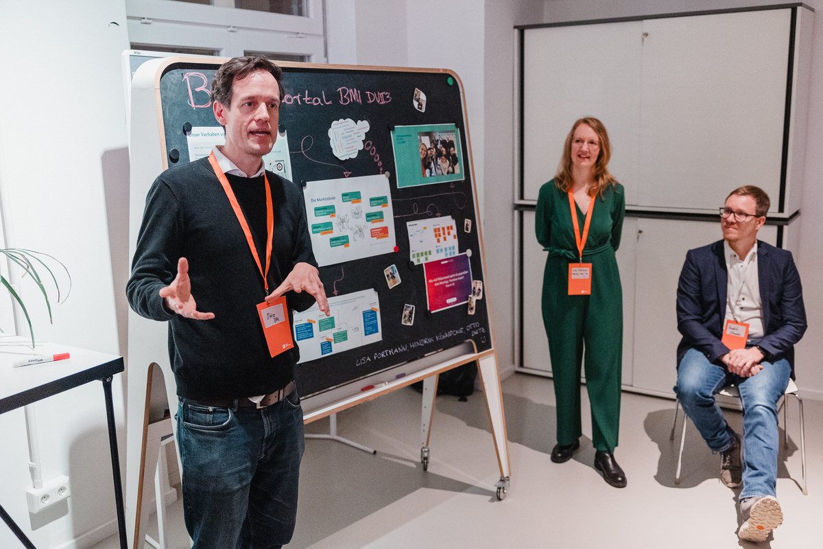 Drei Menschen in einem Büroraum. Projektpartner Otto steht rechts vor einer Tafel und erklärt etwas. Rechts im Hintergrund steht Fellow Lisa und hört zu, neben ihr sitzt Projektpartner Hendrik.