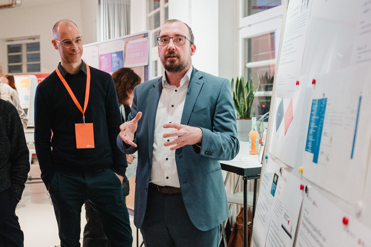 Projektpartner Sascha steht links neben einem Whiteboard mit Notizen und erklärt etwas. Neben ihm steht Fellow Ivo und hört ihm zu. Im Hintergrund sind moderne Büroräume zu sehen.