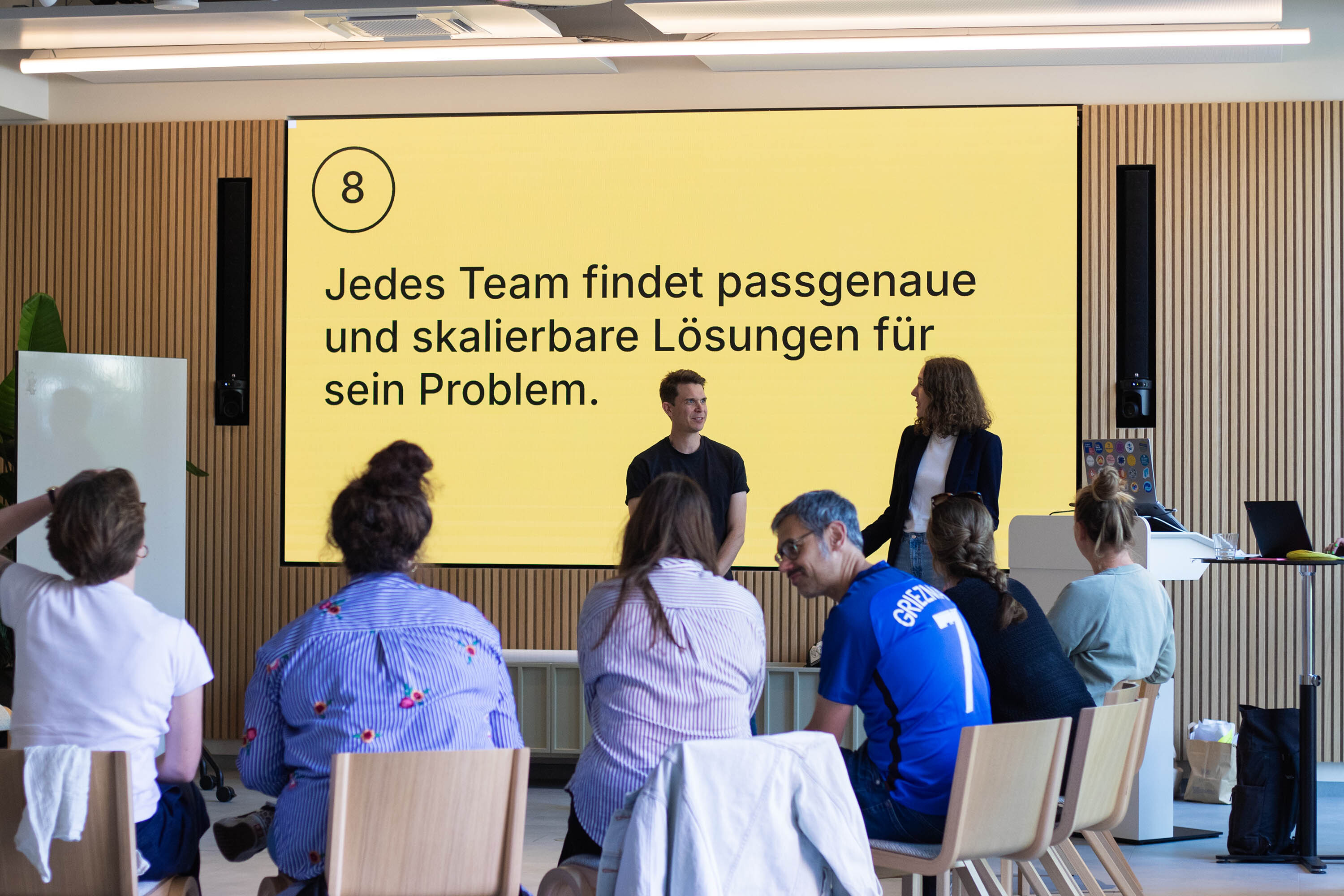 Zwei Personen halten in einem modernen Tagungsraum mit holzvertäfelter Wand und großem Display vor einer Gruppe sitzender Menschen einen Vortrag; auf der Leinwand steht: „8 – Jedes Team findet passgenaue und skalierbare Lösungen für sein Problem.“