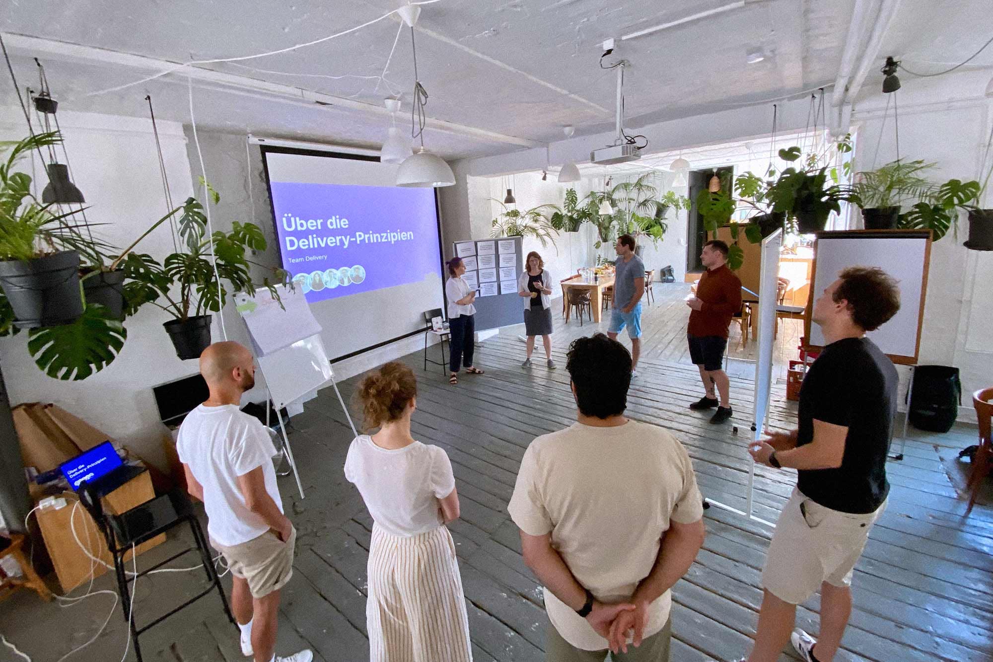 Eine Gruppe von acht Menschen steht kreisförmig in einem Raum und spricht miteinander; an einer Projektionswand ist eine Folie mit der Aufschrift „Über die Delivery-Prinzipien“ zu sehen, und in der Nähe stehen Whiteboards und Flipcharts; die Atmosphäre wirkt ungezwungen und von Zusammenarbeit geprägt