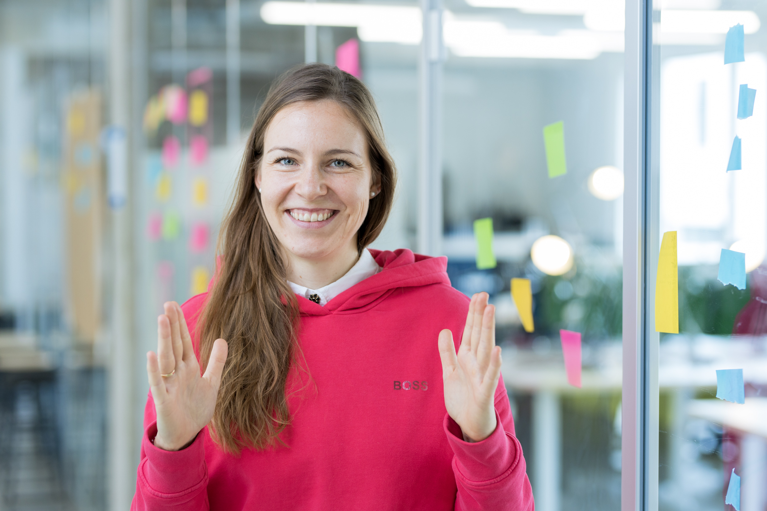 Compliance Managerin Julia macht in einem Büroraum mit Glaswänden voller Klebezettel Gesten mit ihren Händen.