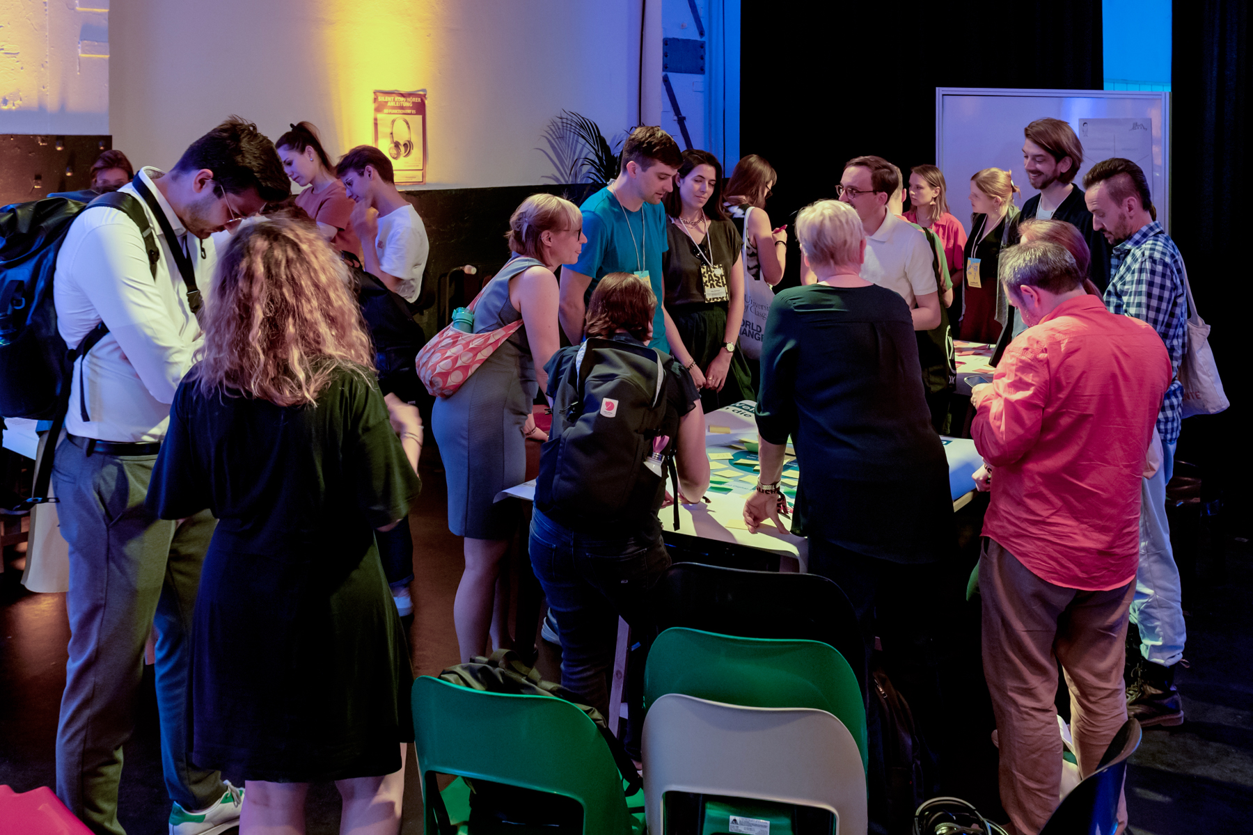 Eine Gruppe vornehmlich junger Menschn beugen sich über einen Tisch, auf dem sich ein Poster befindet, an dem viele Post-its angebracht sind. Links vom Tisch befinden sich weitere Menschen, die sich in kleineren Gruppen unterhalten.