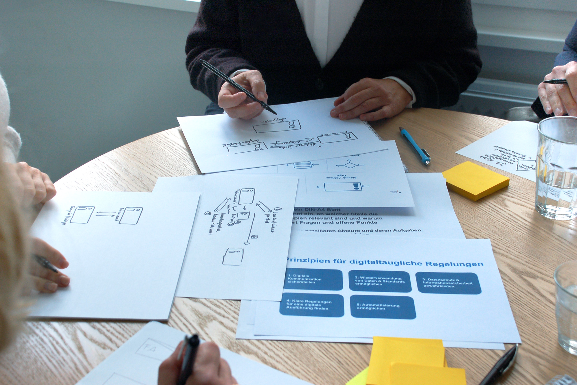 Mehrere Personen sitzen um einen runden Tisch und zeichnen Prozessvisualisierungen auf Papier. Im mittleren Bereich des Tischs liegt ein Papierausdruck mit den fünf Prinzipien für digitaltaugliche Gesetzgebung.