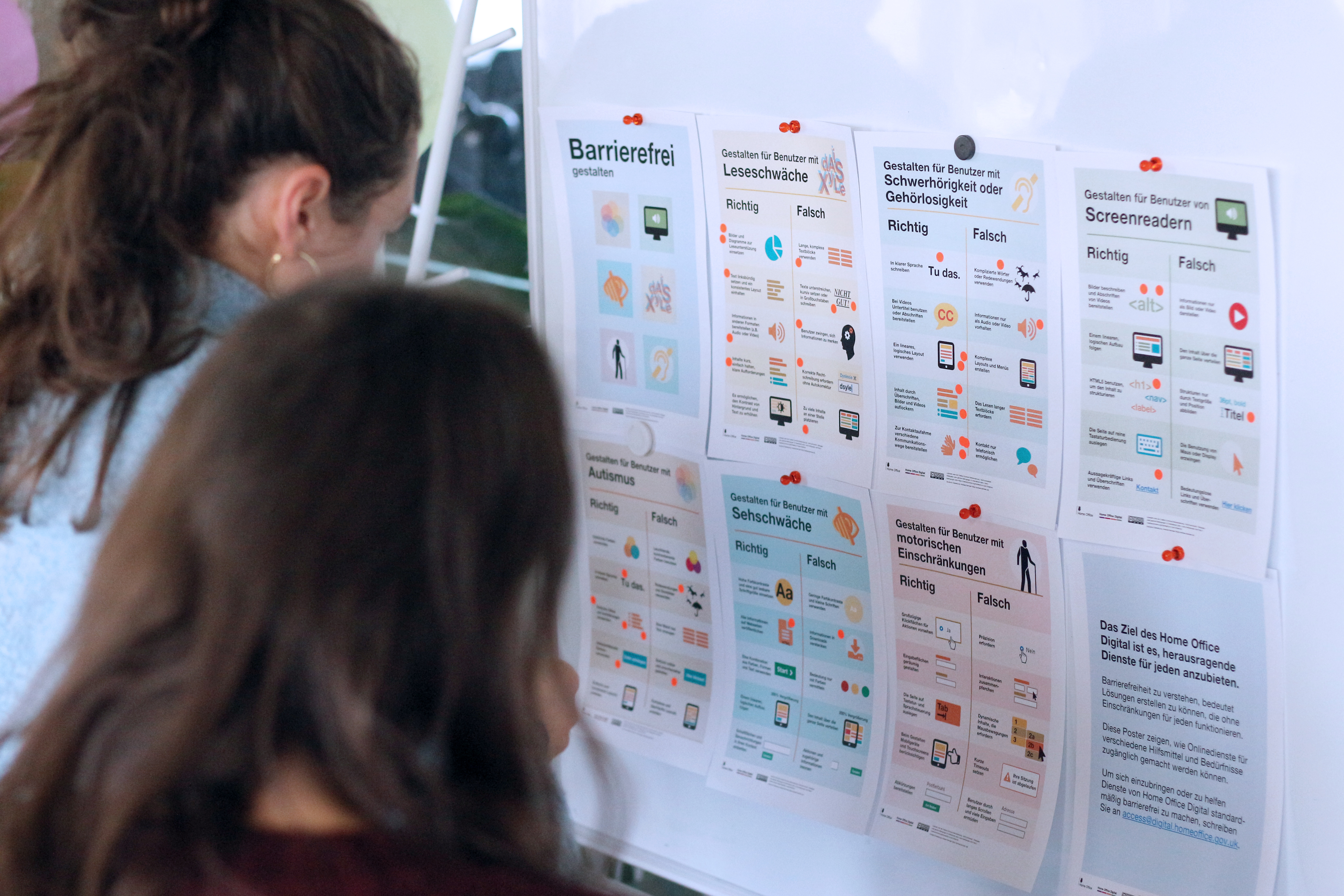 Zwei Personen mit langen Haaren schauen auf 8 kleine Poster zu Barrierefreiheit, die jeweils eine Reihe von 5 Dinge mit Richtig und Falsch überschrieben zeigen
