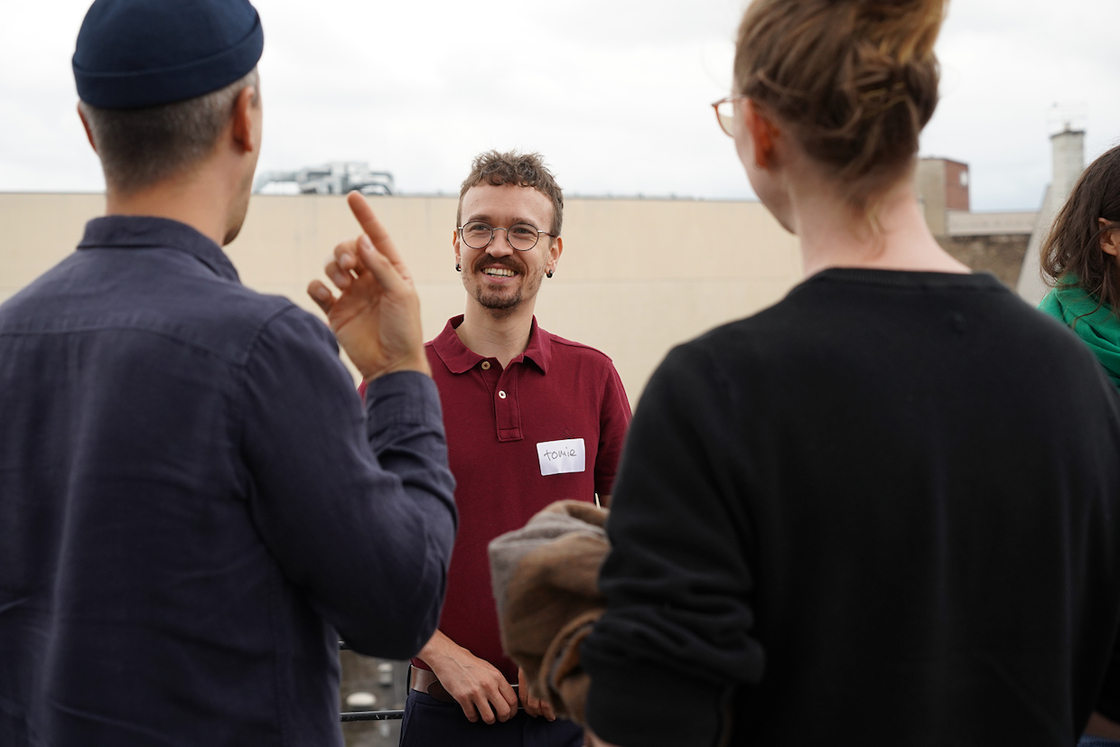 Hier steht Tomie zusammen mit zwei anderen Personen auf der Dachterrasse beim Tech4Germany Auftakt-Event. Einer der Personen ist Tomies Dolmetscher. Der Dolmetscher hilft Tomie in der unterhaltung mit der dritten Person. Tomie hat ein rotes Hemd an, er schaut interessiert in Richtung der der Kamera vorbei.