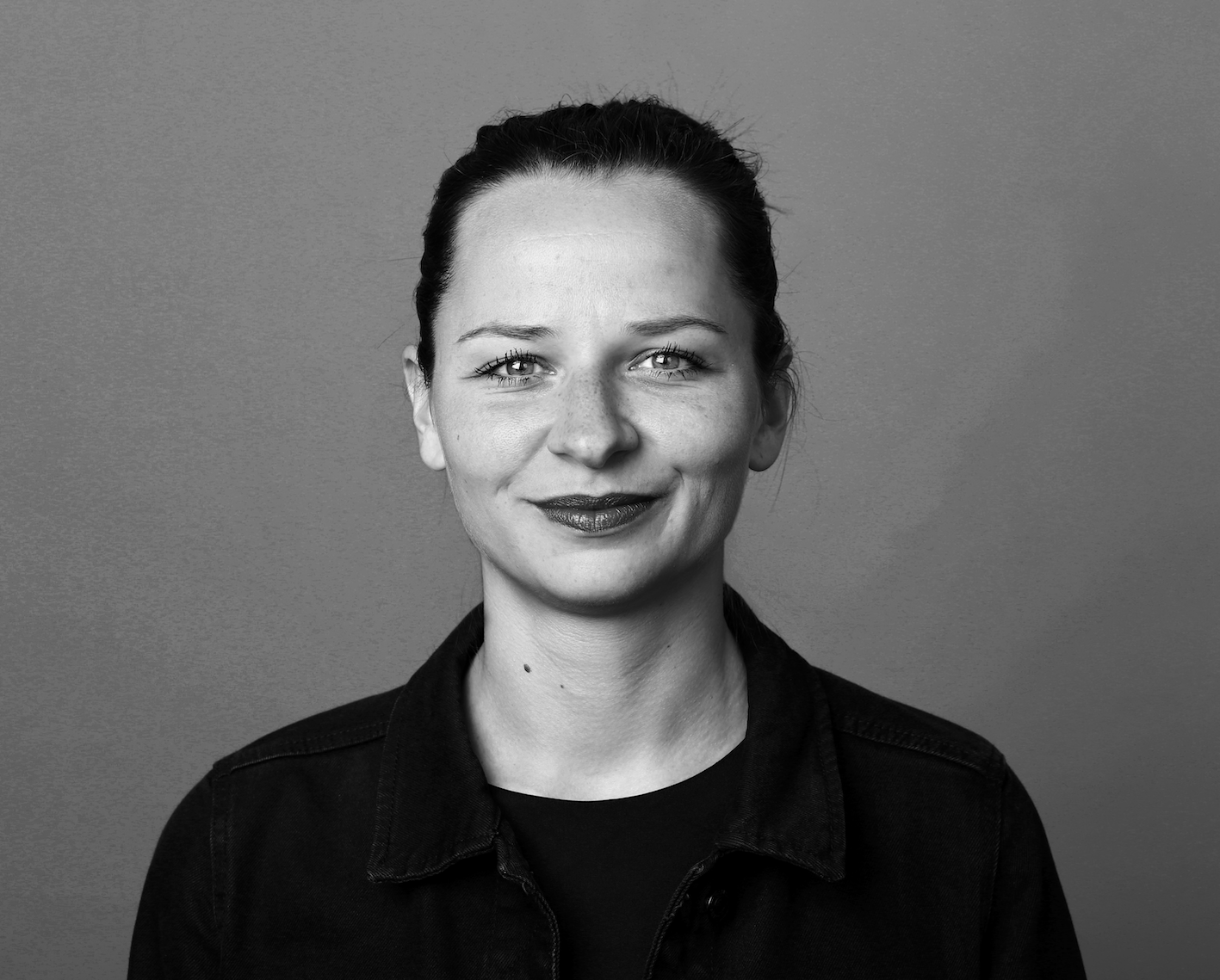 Eine junge Frau lacht in die Kamera; sie trägt ein schwarzes Oberteil und ihre Haare sind zu einem Zopf gebunden; sie steht vor einer grauen Wand; das Portrait ist in Schwarz-Weiß abgebildet.