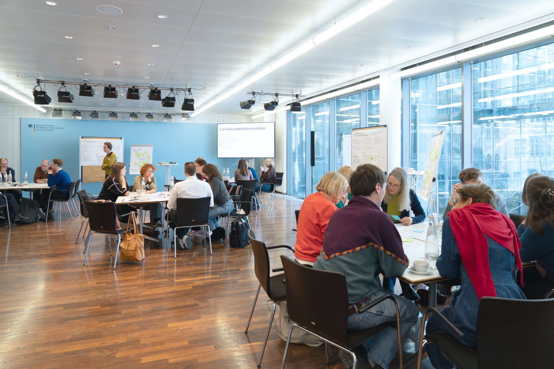 In einem großen Konferenzraum sitzen drei Personengruppen an Tischinseln zusammen und besprechen vor ihnen liegende Dokumente.
