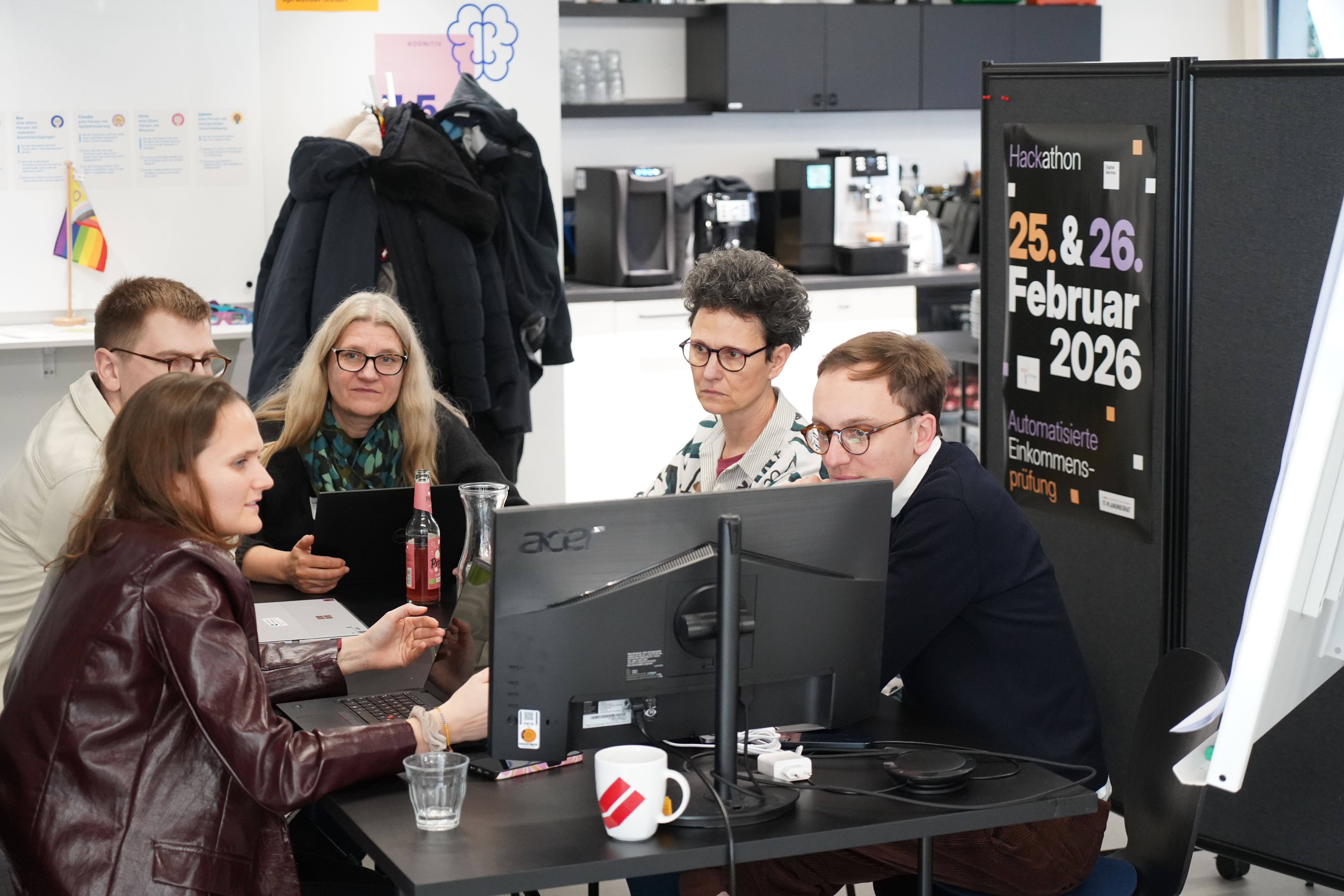 Eine Fünfergruppe sitzt konzentriert im Büro um einen großen Monitor und Laptops herum und bespricht ein Projekt. Im Hintergrund hängt ein Hackathon-Plakat.