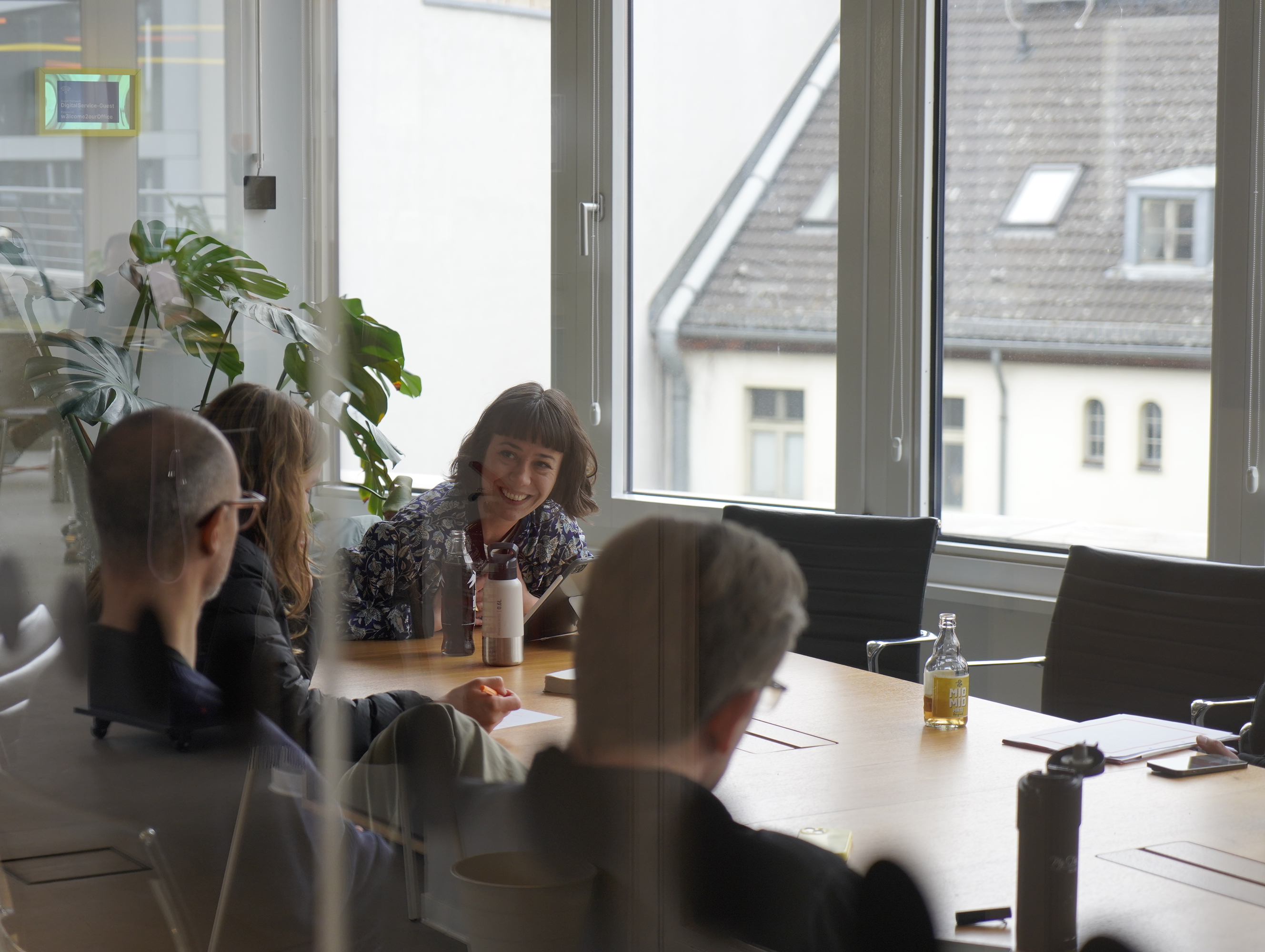 Mehrere Menschen sitzen in einem modernen Büroraum und sprechen miteinander. Im Hintergrund ist das Dach eines Hauses durch eine Fensterfront zu sehen.