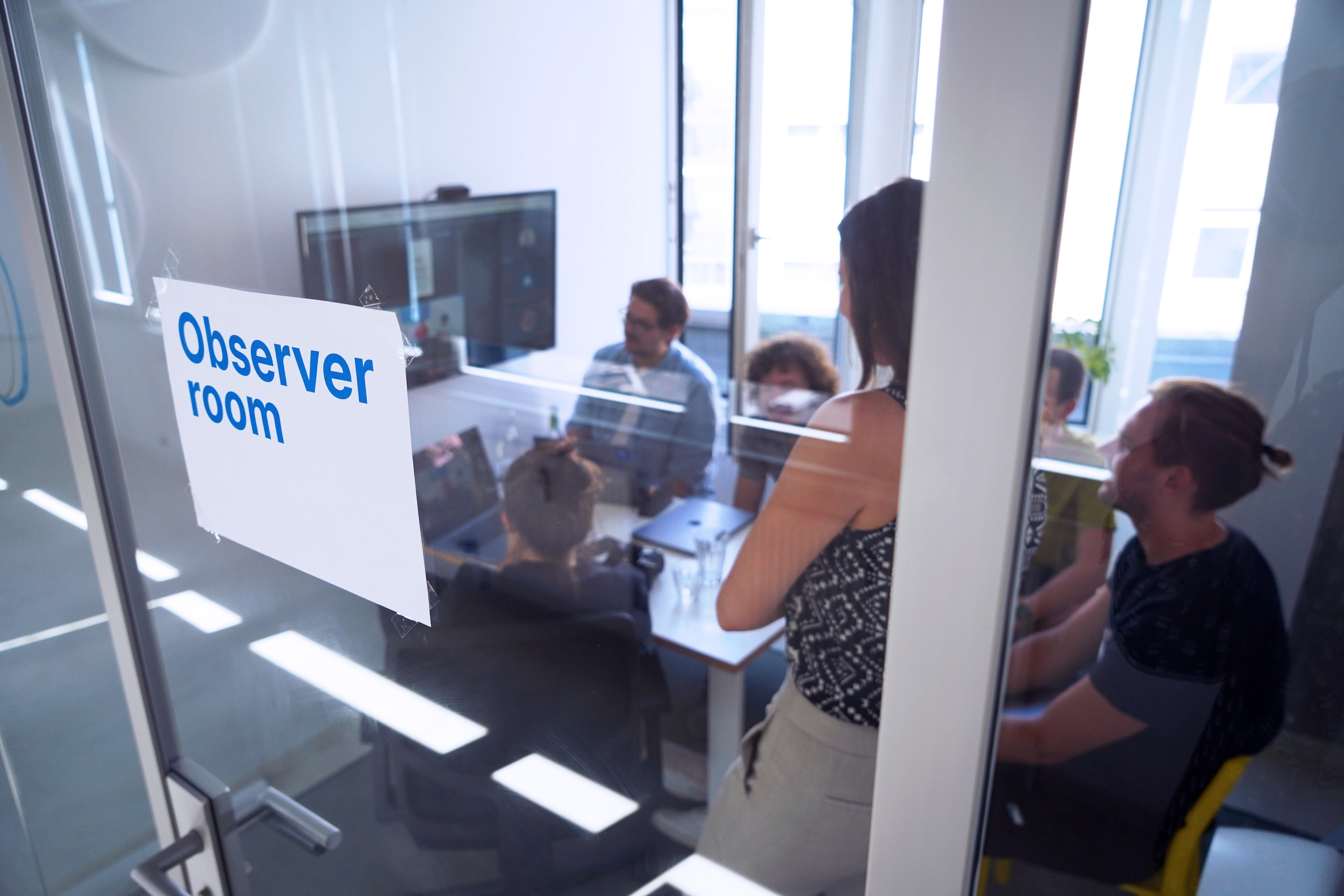 Eine Gruppe von 6 Personen in einem kleinen Raum mit Glaswand, sie schauen alle auf einen Fernseher; an der Glaswand ist ein Blatt Papier befestigt, auf dem steht: Beobachtungsraum auf English.