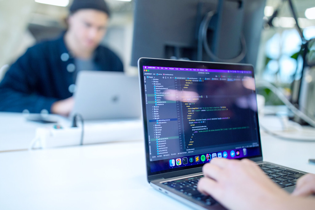 Ein Laptop steht auf einem Schreibtisch. Hinter diesem Laptop ist eine Person unscharf zu erkennen. Auf dem Display des Laptops befindet sich Code.
