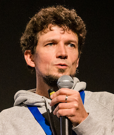 Profilfoto von Fabian Klenk. Er hält ein Mikrofon und präsentiert. Das Foto ist auf dem Abschlussevent der Fellowships im Herbst 2022 entstanden.