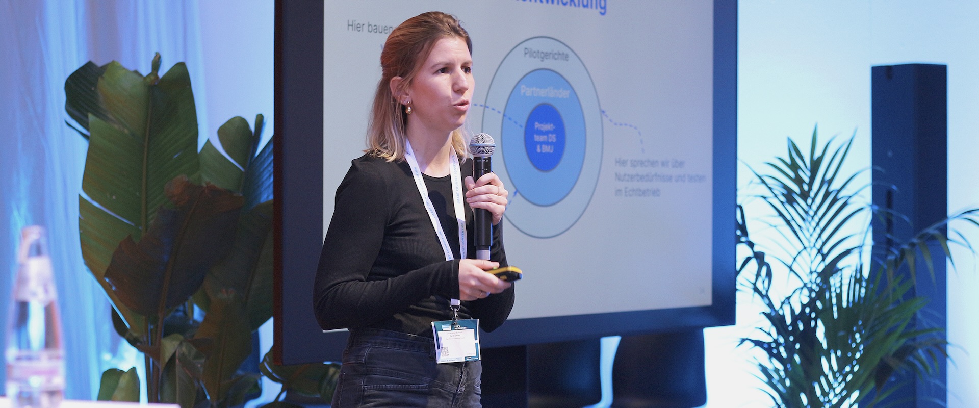 Die Autorin Caroline hält ein Mikrofon in der Hand und steht vor einem Bildschirm auf einer kleinen Konferenzbühne.