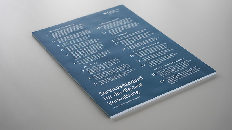 Auf einer weißen Tischplatte liegt ein Stapel mit Postern, auf denen die 19 Punkte des Servicestandards für die digitale Verwaltung beschrieben sind.