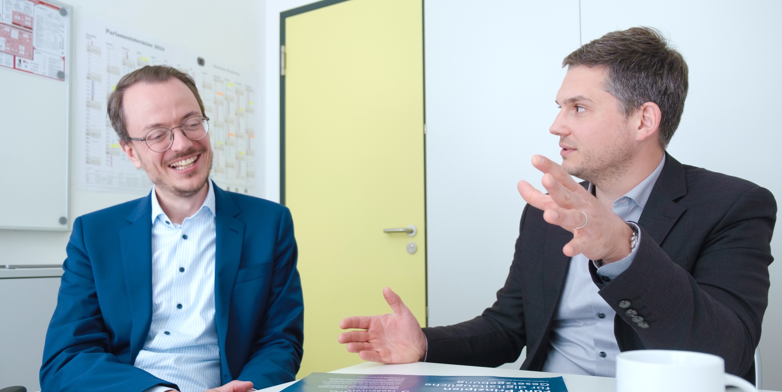 Zwei Männer in einem Gespräch, einer lächelnd in einem blauen Anzug, der andere gestikulierend in einem dunklen Sakko. Im Hintergrund ein Türrahmen und Pinnwände.