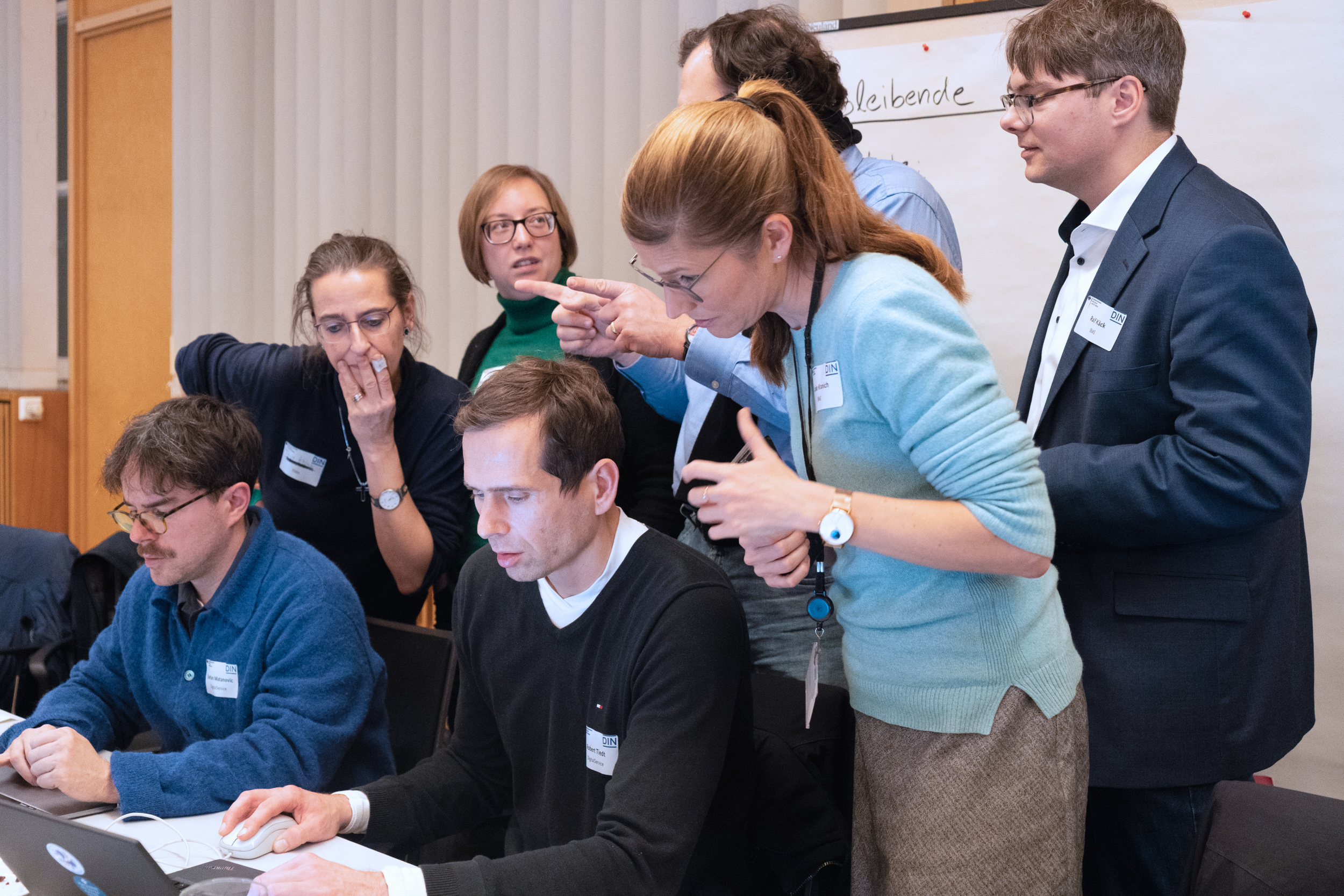 Eine Gruppe von sieben Mitgliedern des Konsortiums zur Erarbeitung der DIN SPEC 66336 diskutiert angeregt. Zwei der Personen sitzen und arbeiten am Computer, während fünf Personen stehend auf den Bildschirm zeigen und gestikulieren. Im Hintergrund ist ein Whiteboard mit Notizen zu sehen.